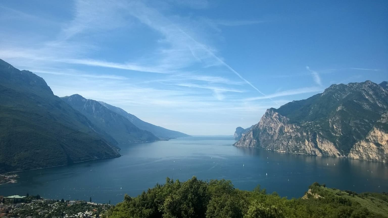 The northern end of Lake Garda seen from a green hillside, the lake running into the distance between steep mountain walls, small sailboats on the water and a lakeside town in the lower left corner