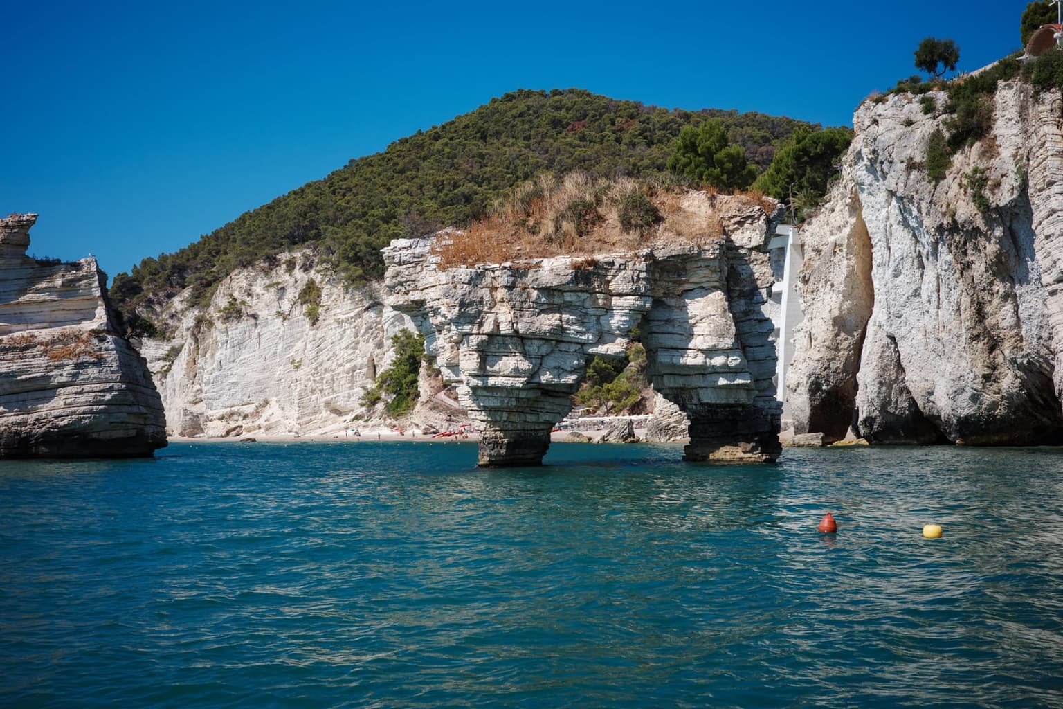 The limestone arch stack at Baia delle Zagare seen from a boat, with the small beach and a white stairway behind it