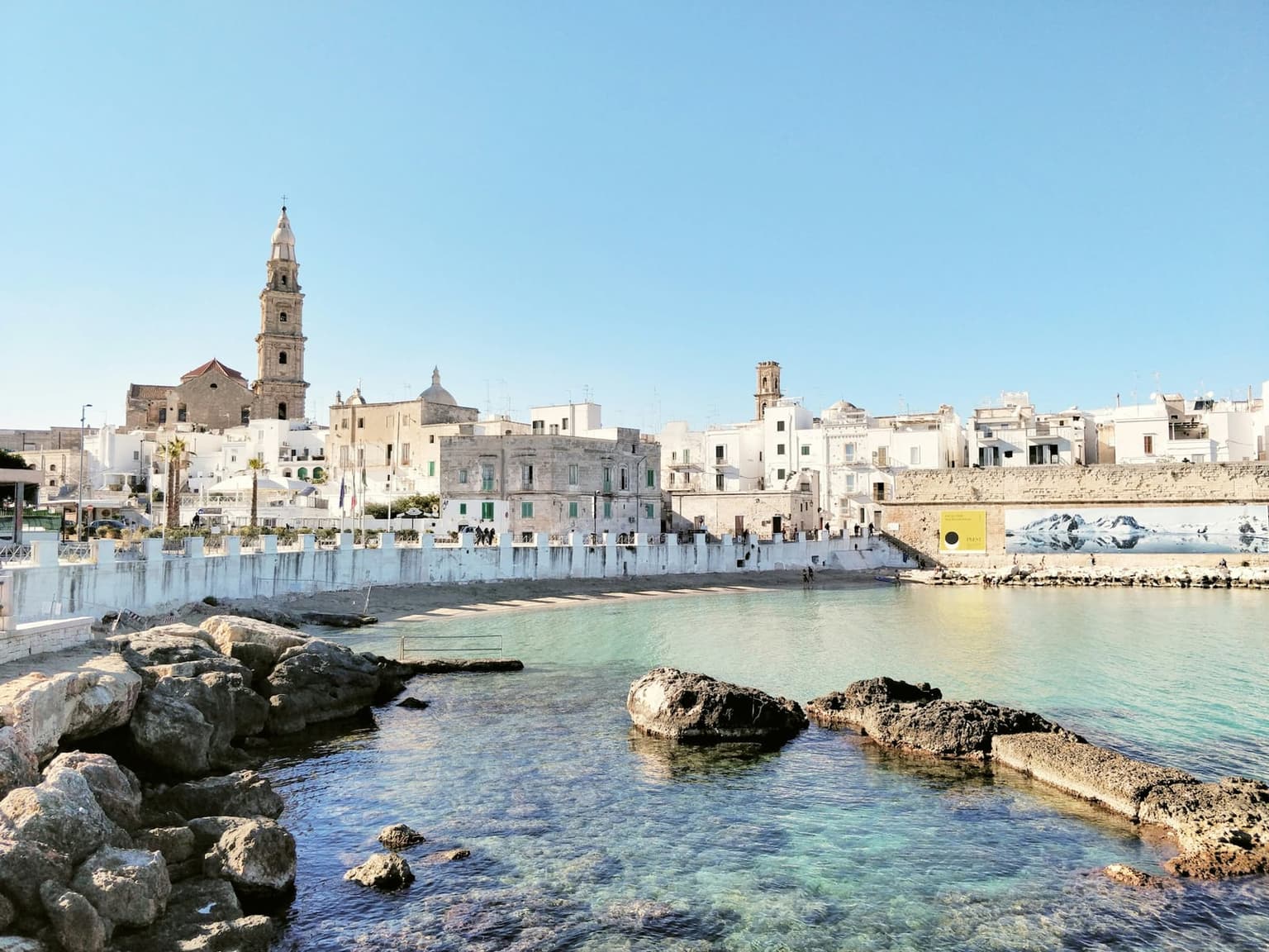 The old town sea wall and cathedral bell tower above the small rocky cove of Cala Porta Vecchia in Monopoli, with clear shallow water in front