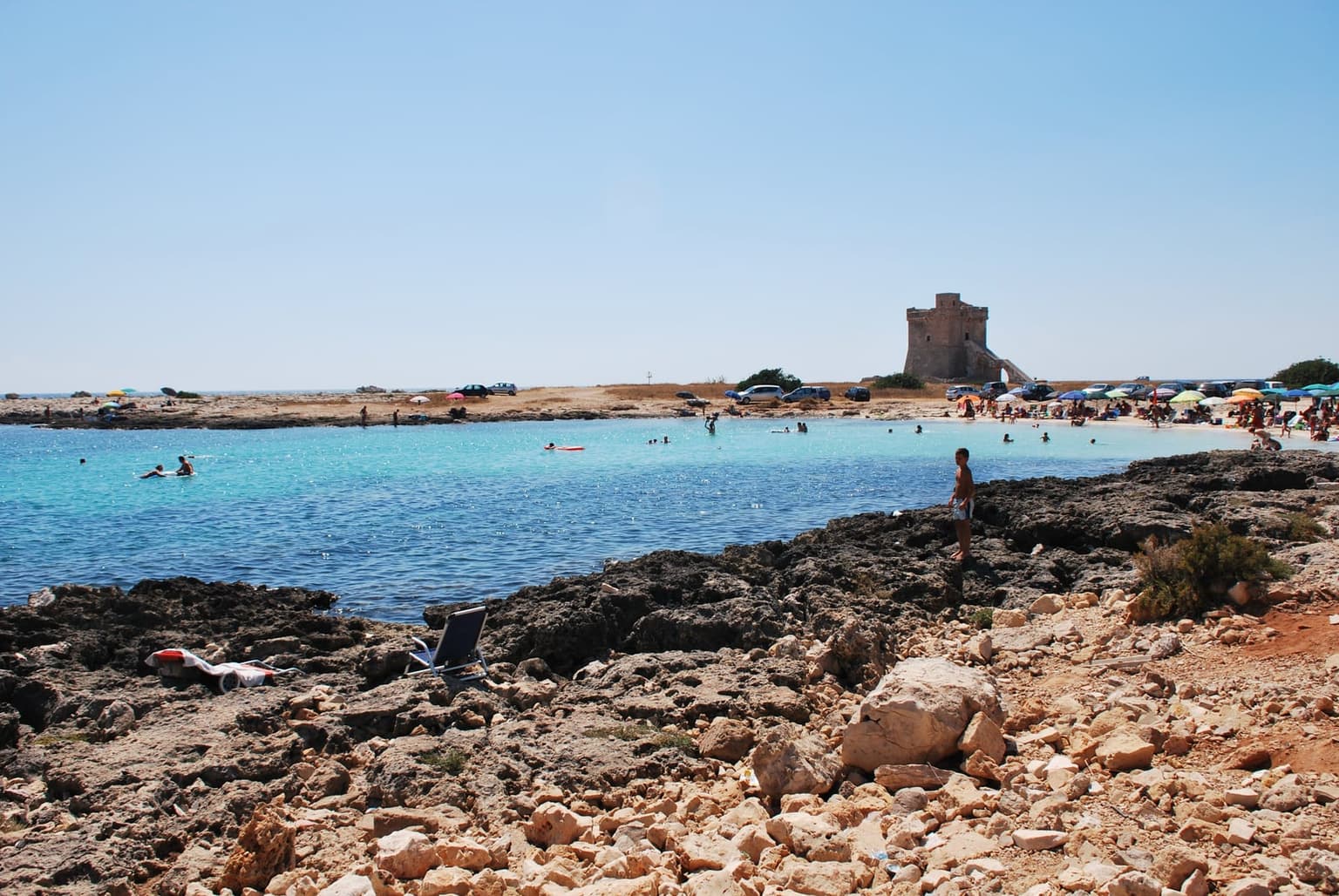 A calm turquoise cove on the Ionian side near Porto Cesareo, with a watchtower on the point and swimmers in the shallows