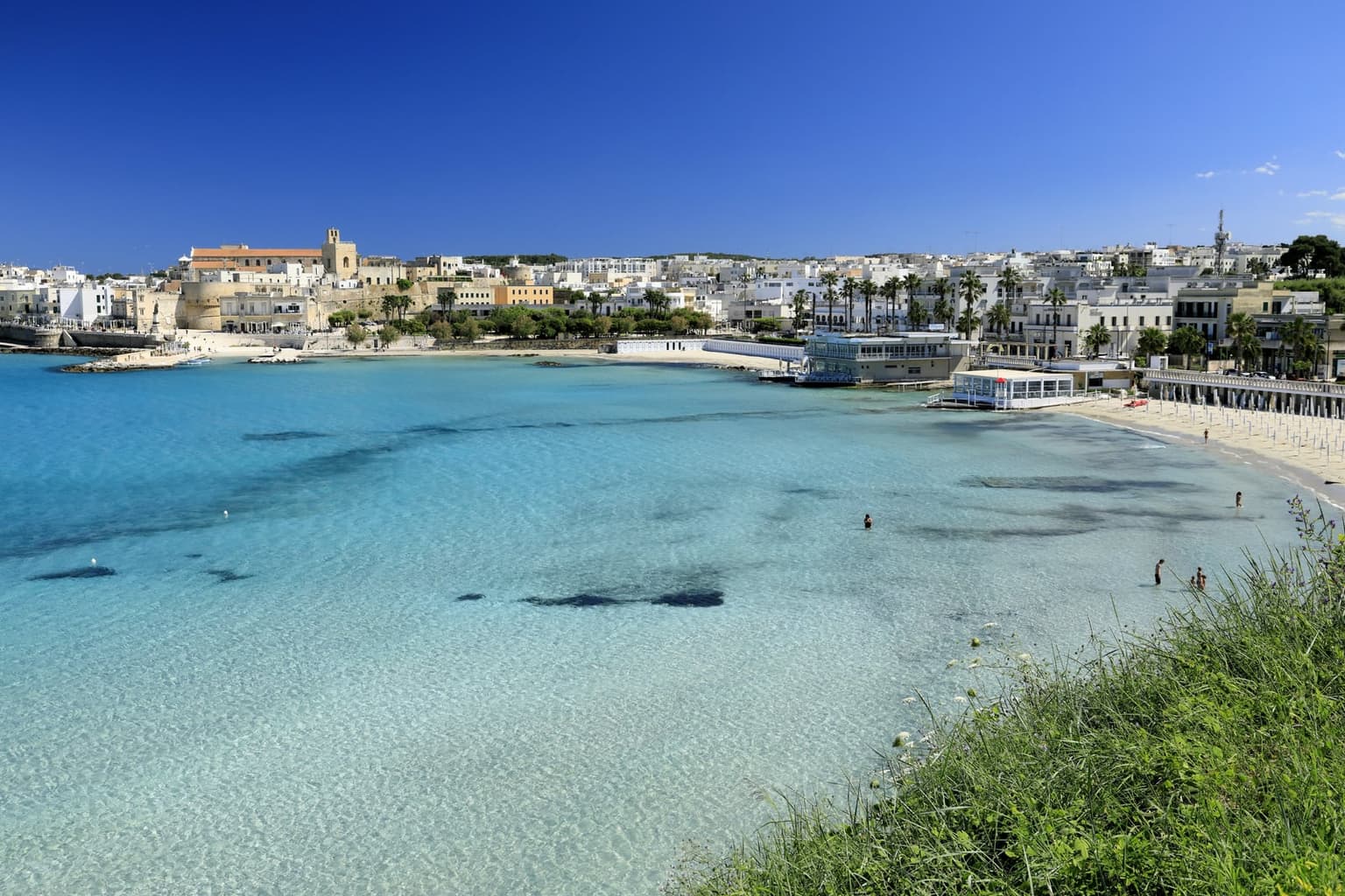 Otranto's town beach seen from the cliff path, with turquoise shallows, a sand crescent, and the old town with its castle walls and cathedral behind
