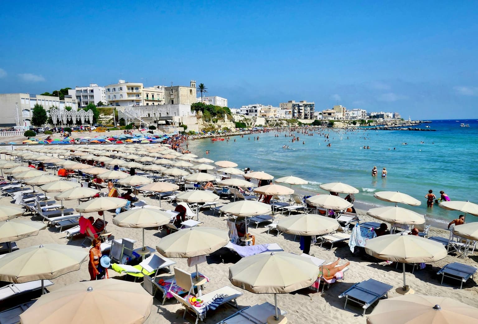 Rows of parasols and sunbeds at a lido on the town beach in Otranto, with the old town behind