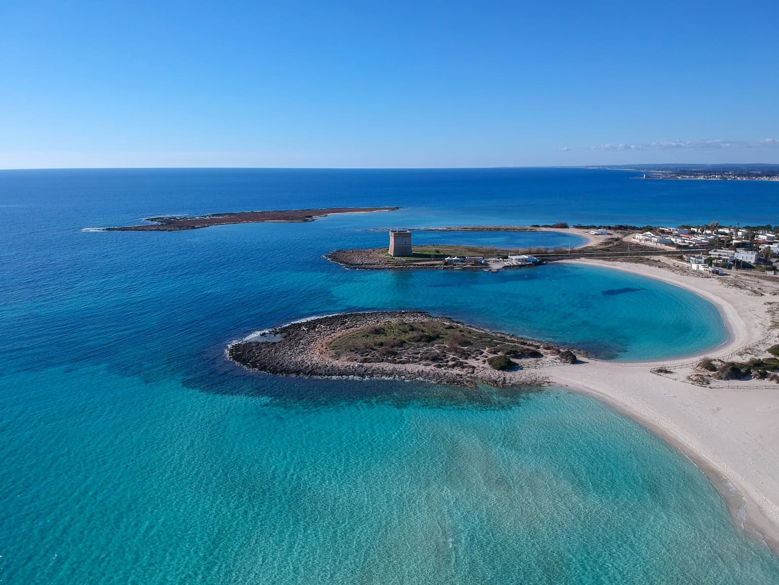 Low aerial view of the Porto Cesareo coast on the Ionian side of the Salento: a pale sand crescent with a watchtower and small islets in shallow turquoise water
