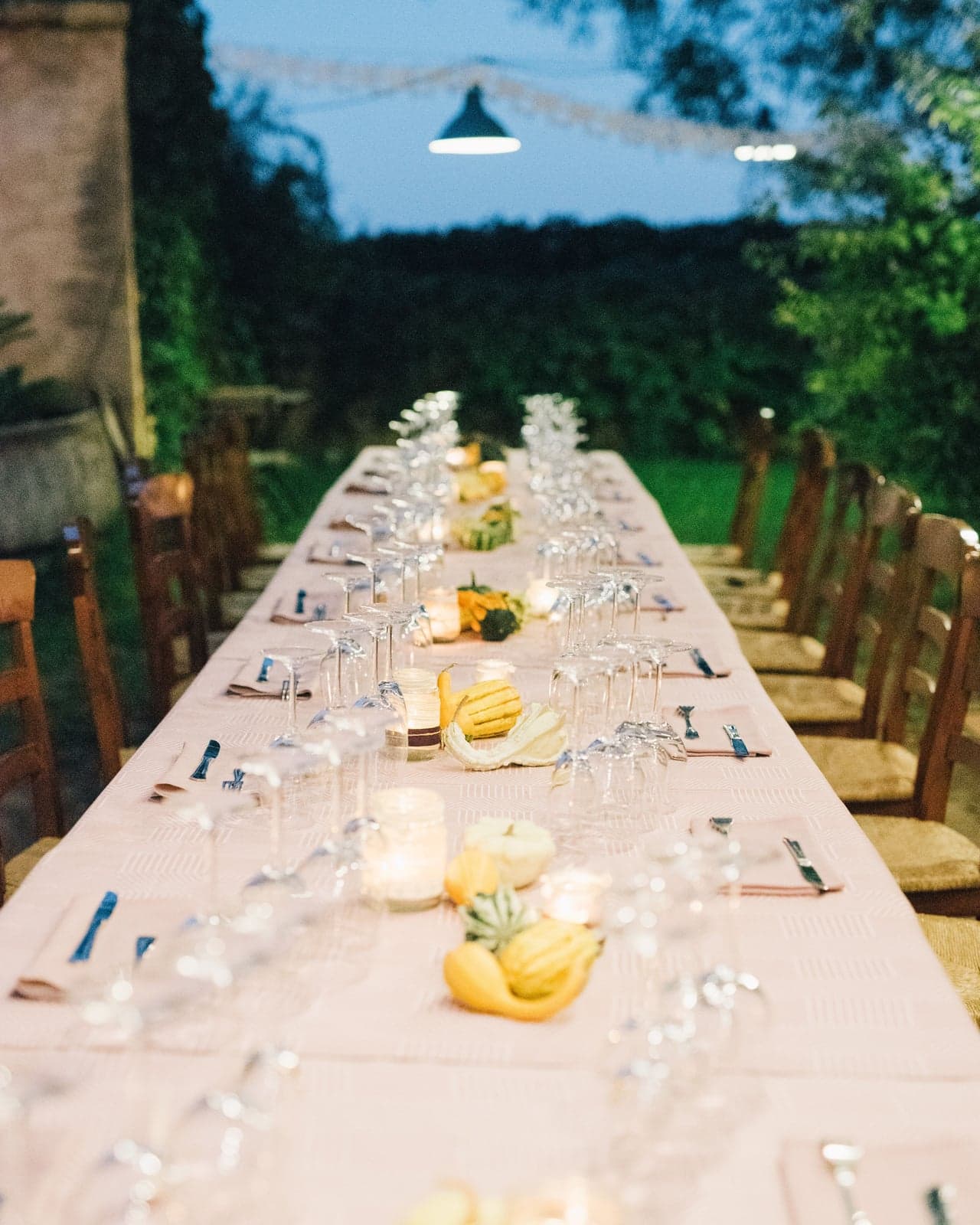 A long table laid outdoors for dinner at dusk, candles in glass jars and small gourds down the middle, wine glasses and folded napkins at every place, with string lights overhead and a stone wall and trees behind