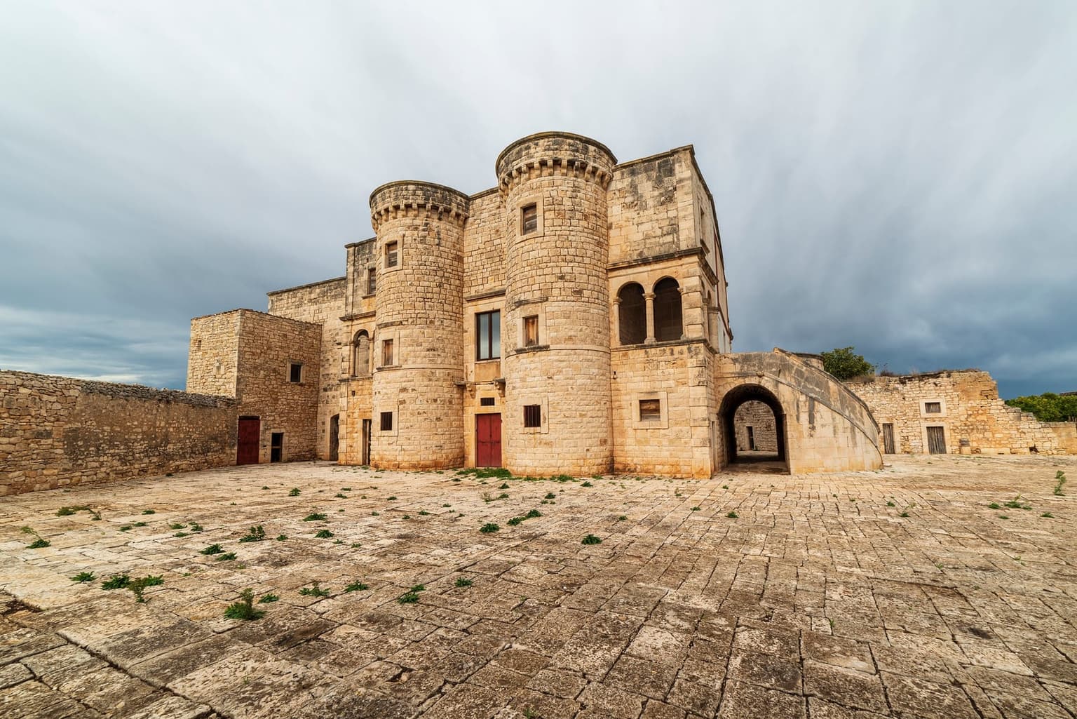 A fortified stone farmhouse with two round crenellated towers and a twin-arched loggia, across an empty stone-paved courtyard with long rubble walls on either side
