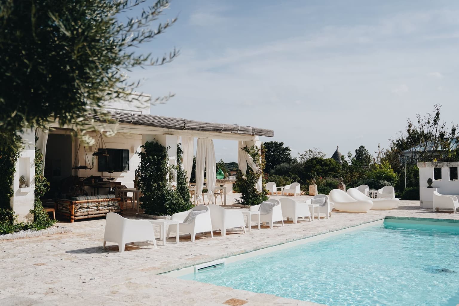 A pool on a stone terrace beside a whitewashed building with a reed-covered pergola, white loungers along its edge and the cone of a trullo roof among the trees behind