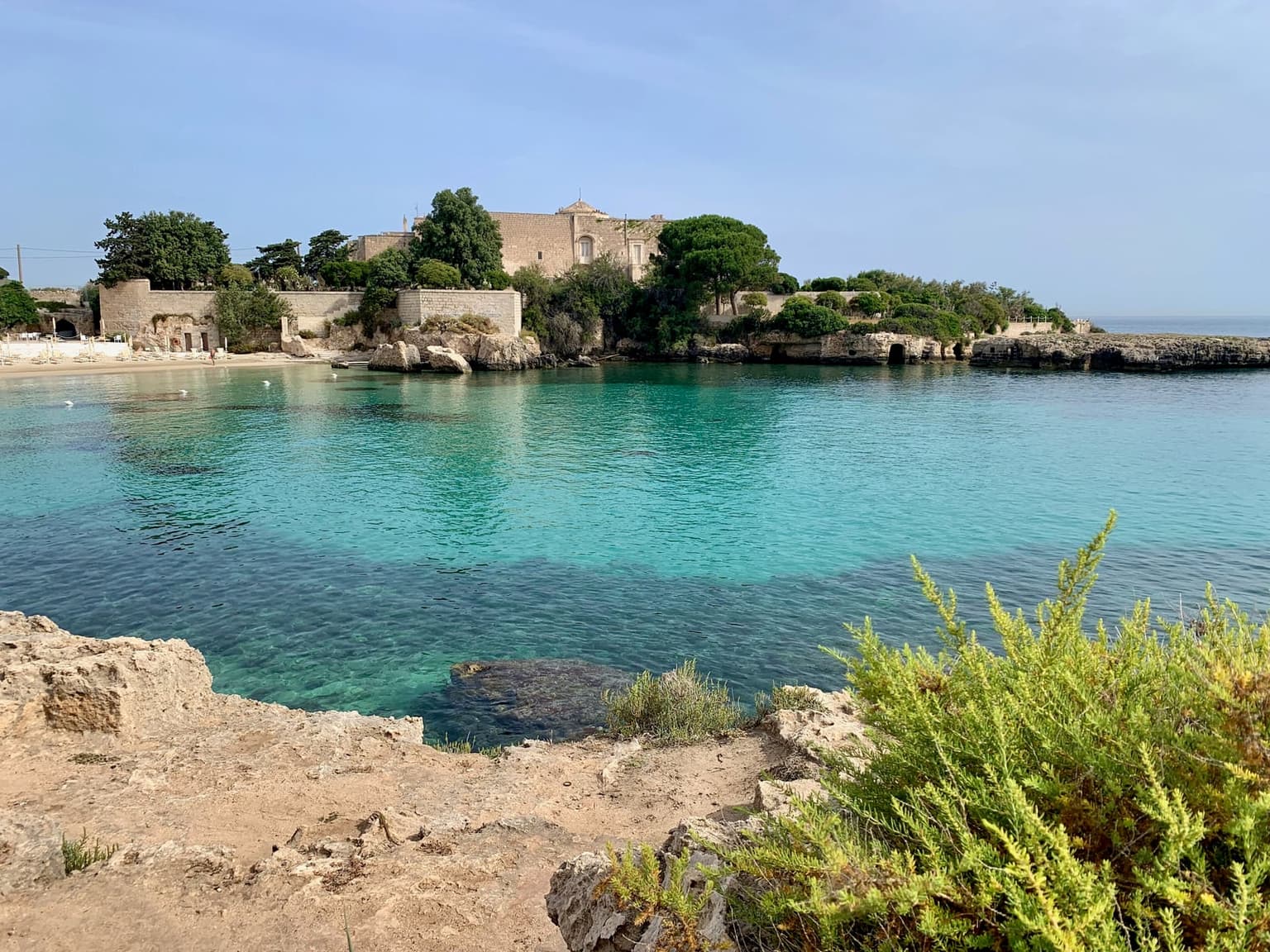 Santo Stefano Castle, a former abbey, among pine trees on a low rocky peninsula above a clear turquoise cove near Monopoli, with a sand beach and sunbeds at the far left and limestone rocks in the foreground