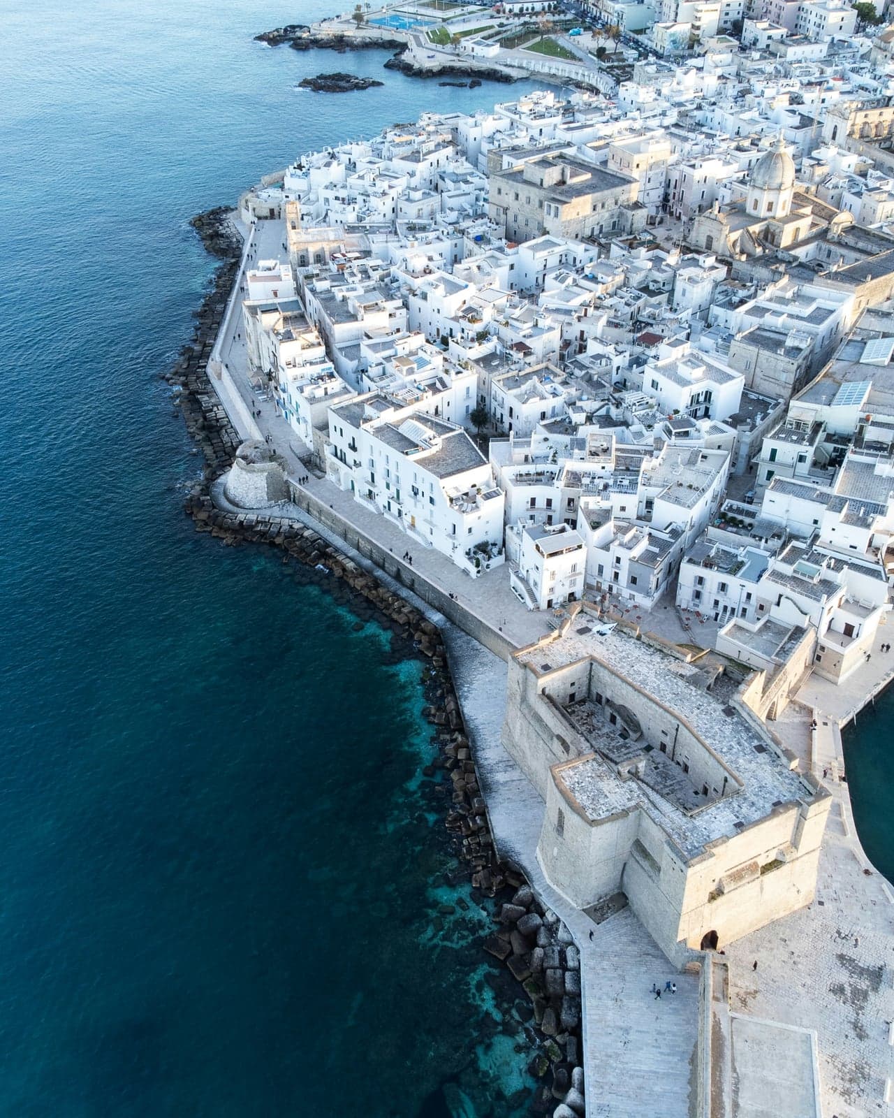 Aerial view of the Castle of Charles V in Monopoli, a square stone fort with an open inner courtyard at the end of the old town's sea wall, deep blue water along one side and the whitewashed houses of the old town behind