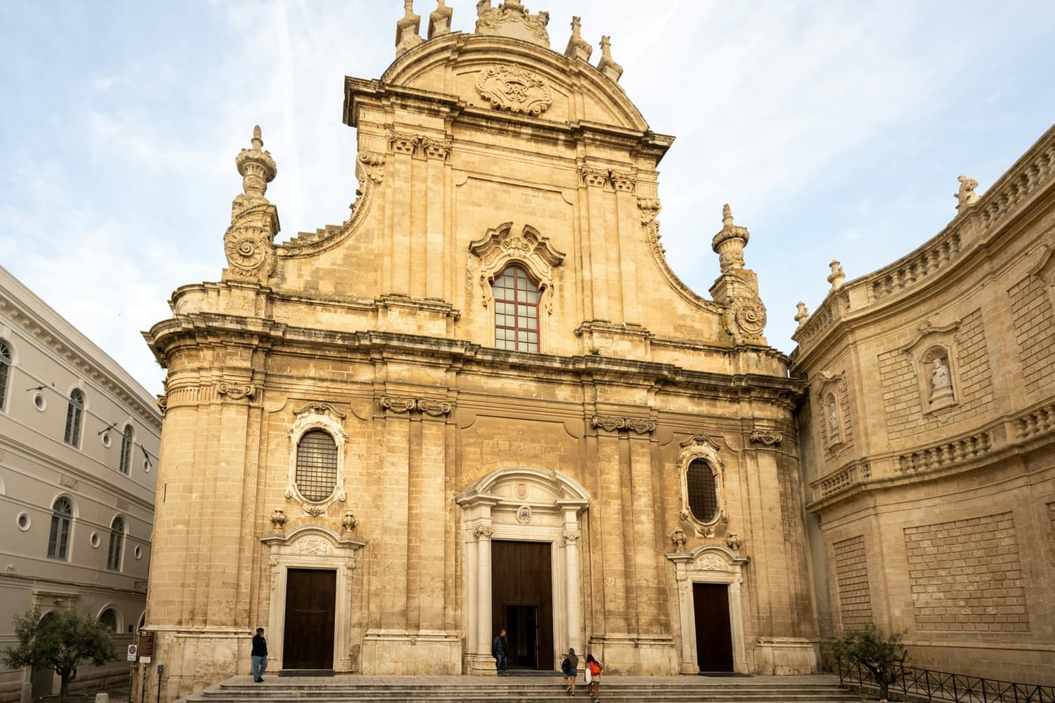 The Baroque limestone facade of Monopoli cathedral, two tiers with a scrolled pediment, a carved crest and spire finials above three wooden doors, a few visitors on the front steps and a curved wing with statues in niches to the right