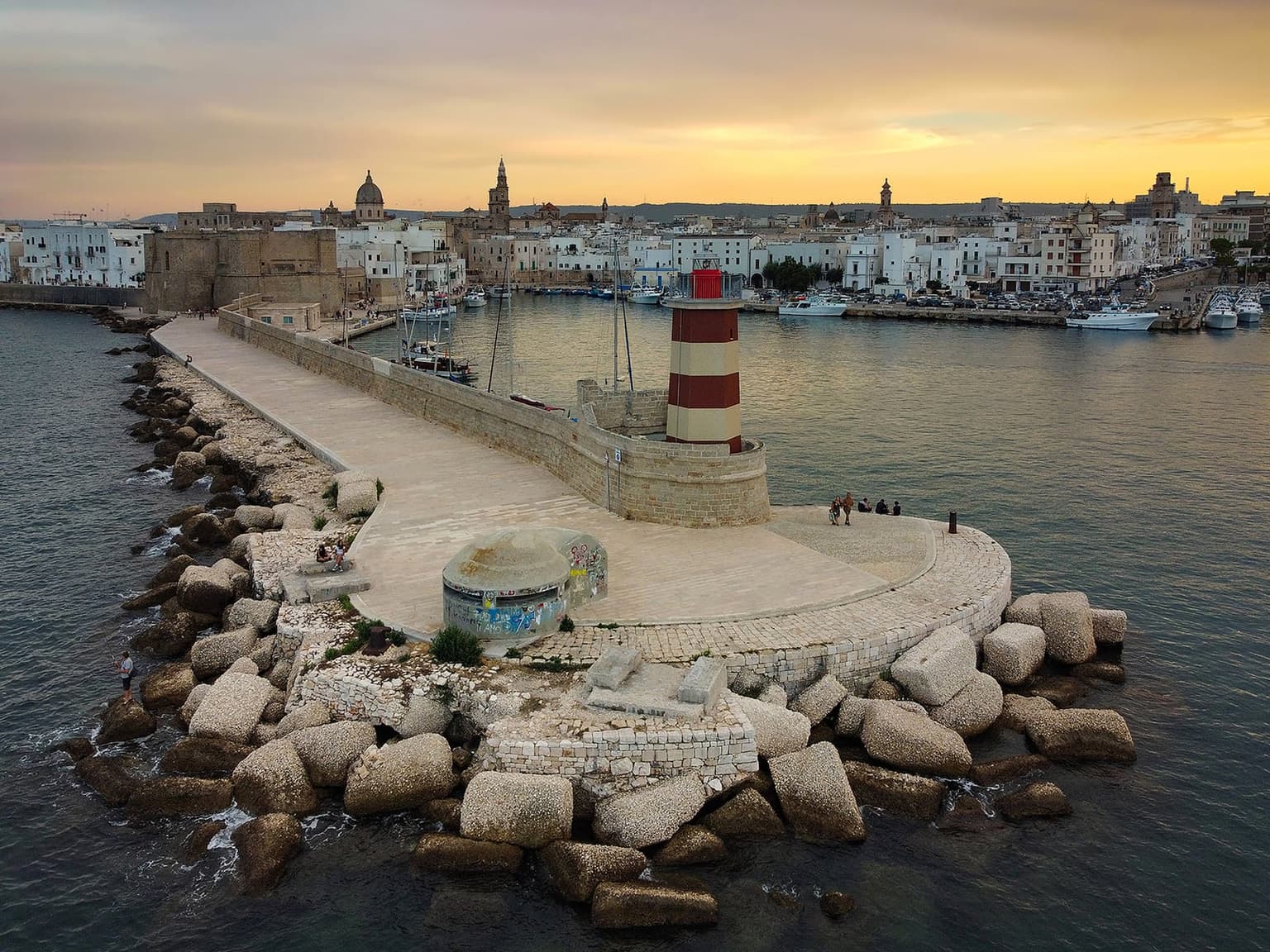 The harbor breakwater in Monopoli at sunset, a red-and-white striped lighthouse at the end of the pier with a few people sitting on its edge, and across the calm harbor water the old town with its castle, a bell tower and a dome under a golden sky