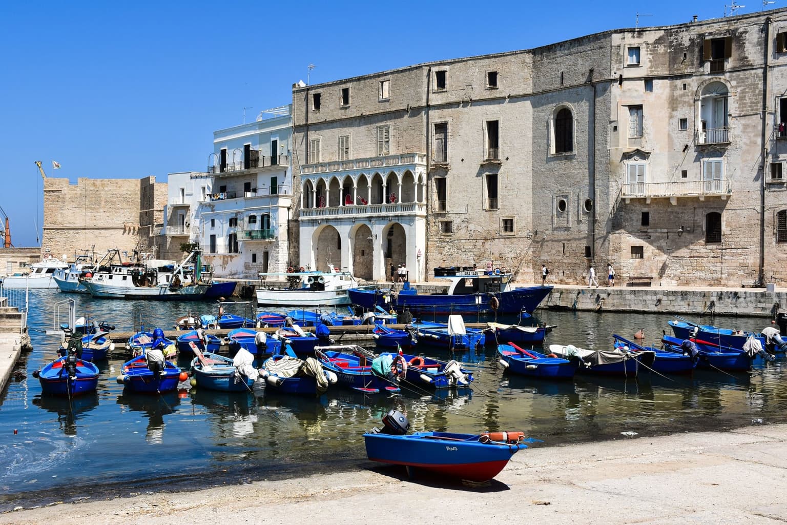 Small wooden fishing boats moored in Monopoli's old harbor below weathered stone buildings and a palazzo with an arched white loggia, larger fishing boats at the quay behind, part of the castle walls at the left and one boat pulled up on the slipway in the foreground