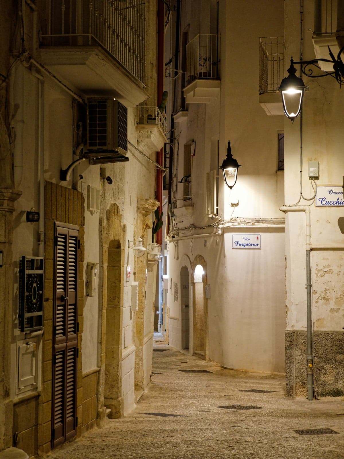 A narrow stone-paved lane in Monopoli's old town at night, lit by wrought-iron lanterns, with balconies and stone doorways along whitewashed walls and a street sign reading Vico Purgatorio