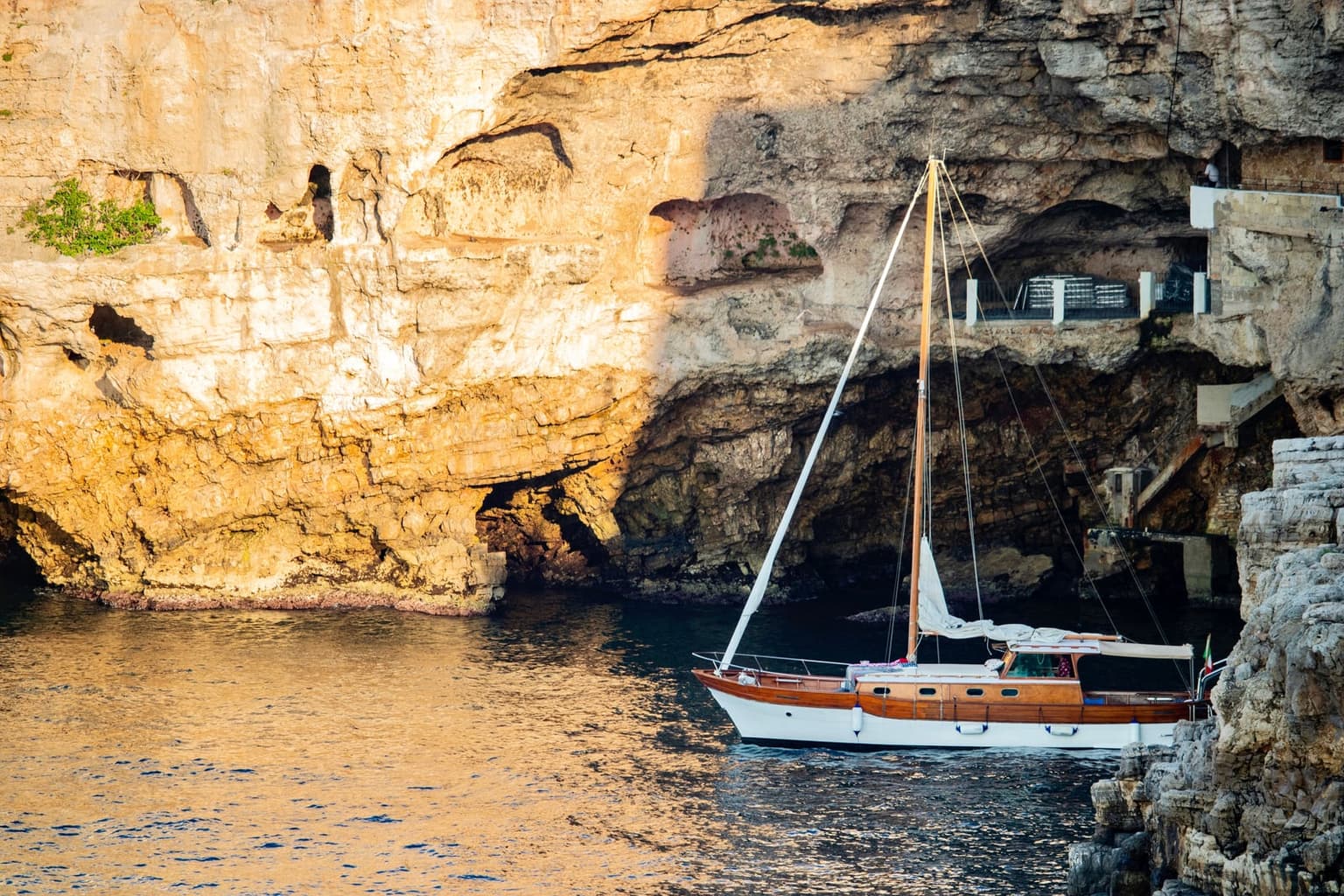 A wooden sailboat passing a large sea cave in the sunlit limestone cliffs at Polignano a Mare, with smaller cave openings along the waterline and a railed terrace built into the cave mouth