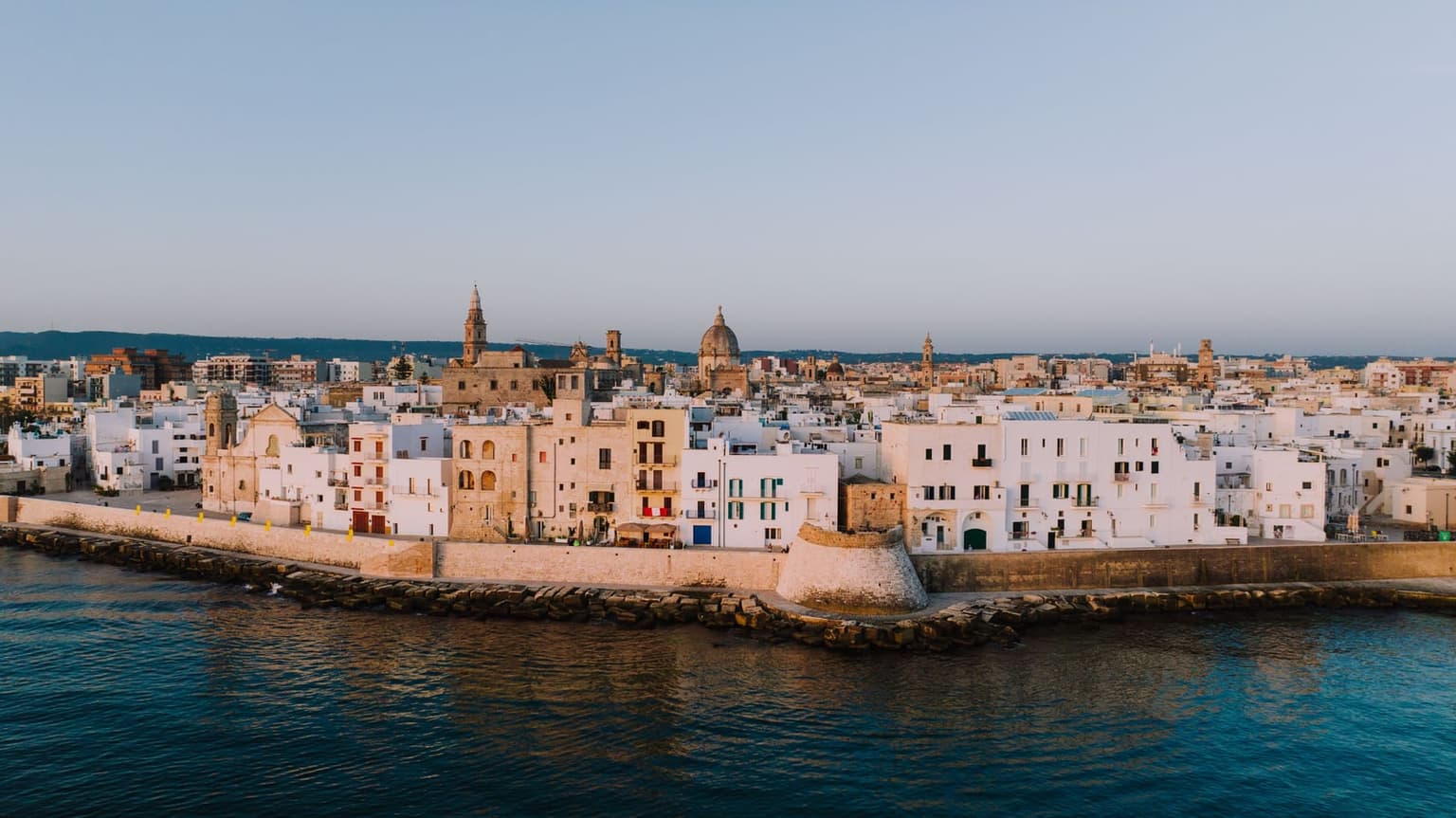Monopoli's old town seen across calm water at golden hour: a stone sea wall with a round bastion along the shore, whitewashed and honey-colored houses behind it, and a bell tower and a church dome above the rooftops