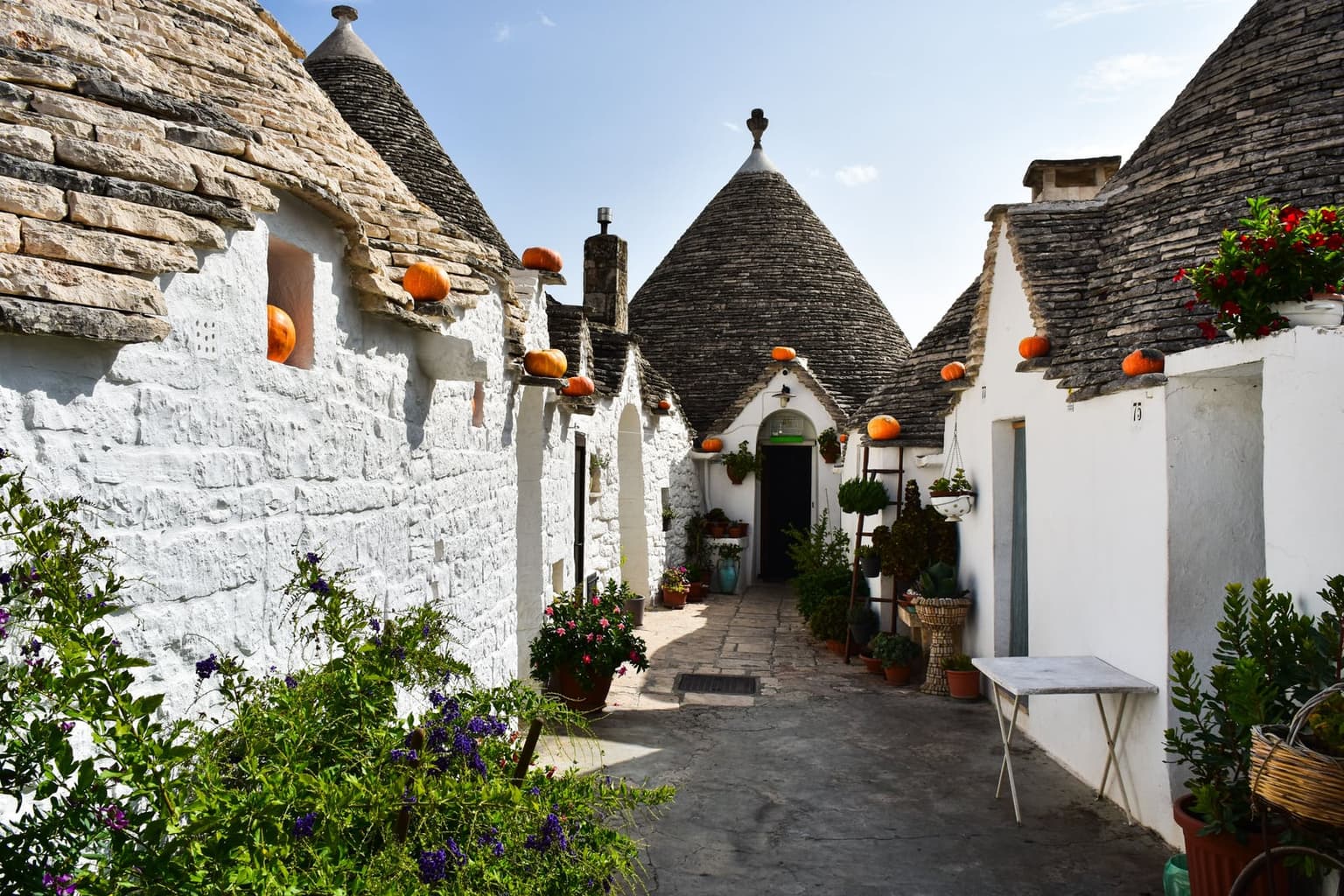 A narrow paved lane between whitewashed trulli with gray conical stone roofs in Alberobello, with potted plants and small pumpkins on the eaves.