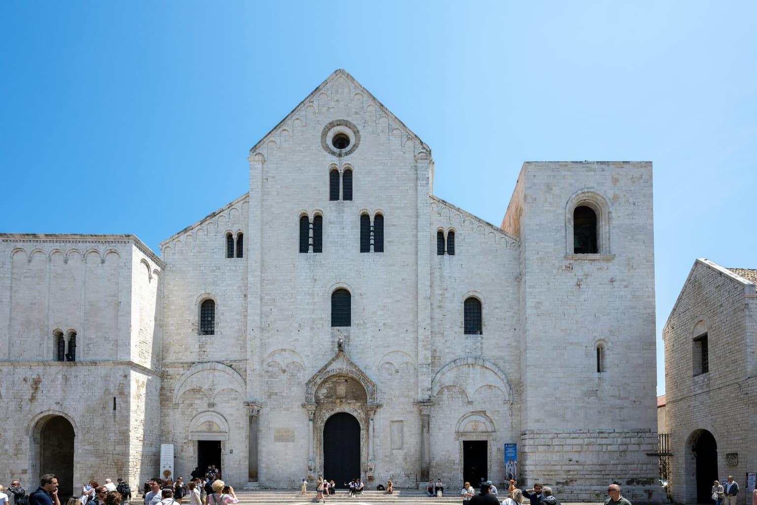 The pale limestone Romanesque facade of the Basilica di San Nicola in Bari Vecchia, with a gabled front, a square tower and visitors on the steps.
