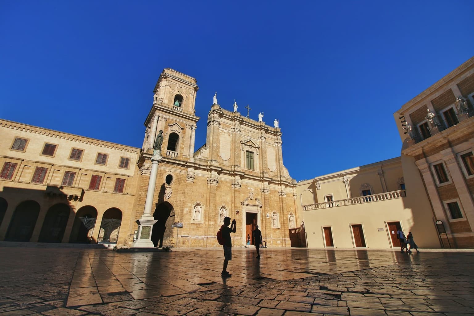 Baroque cathedral facade with a bell tower and a tall column in a sunlit, stone-paved square in Brindisi, under a deep blue sky.