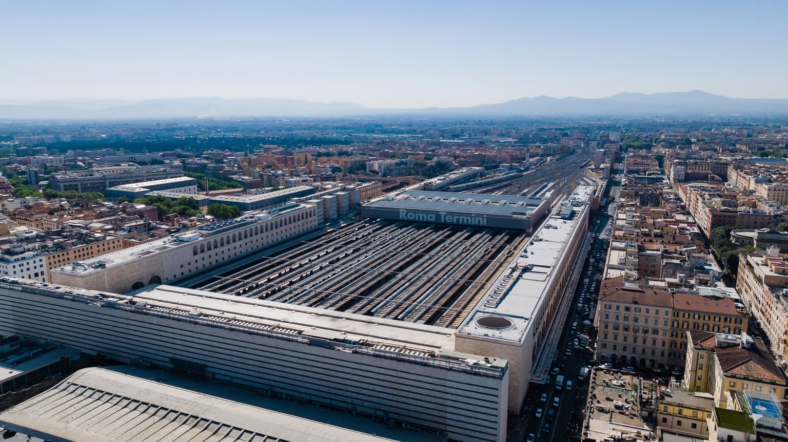 Aerial view of Roma Termini on a clear day, with the station name on the roof of the train shed and the long platform roofs running away between the surrounding blocks.