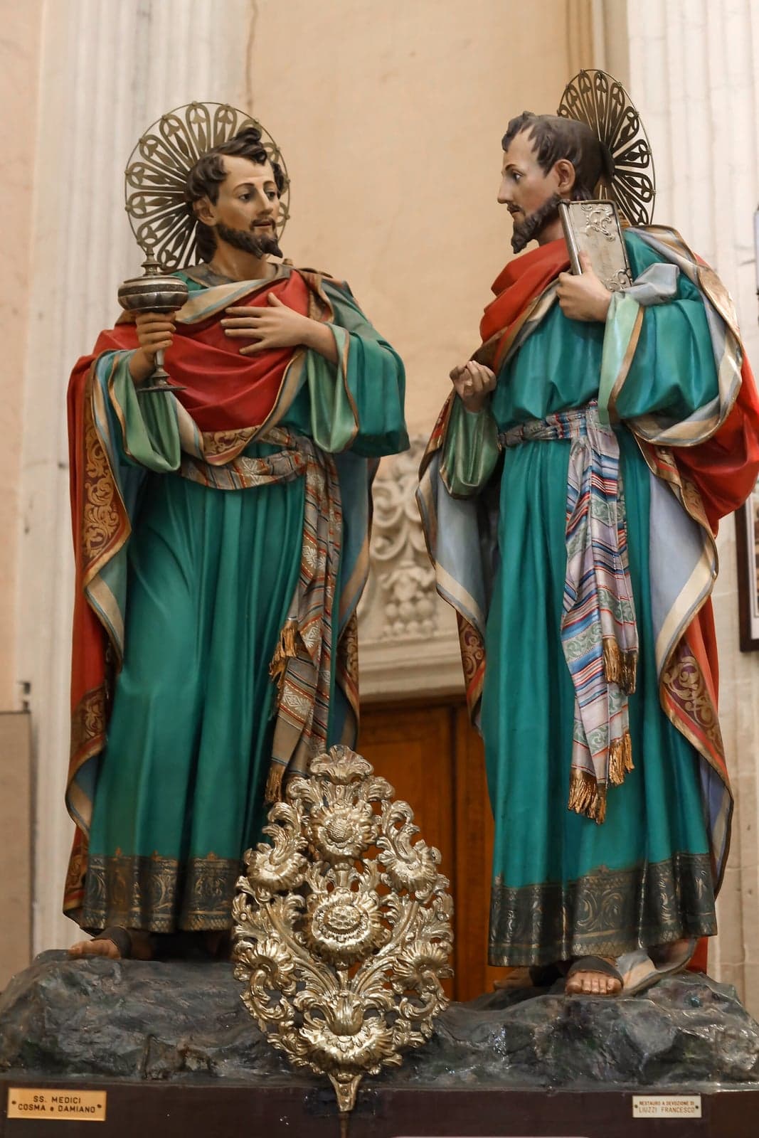 Painted statues of Saints Cosmas and Damian in a Lecce church, in green robes and red cloaks with metal halos, one holding a lidded vessel and the other a book
