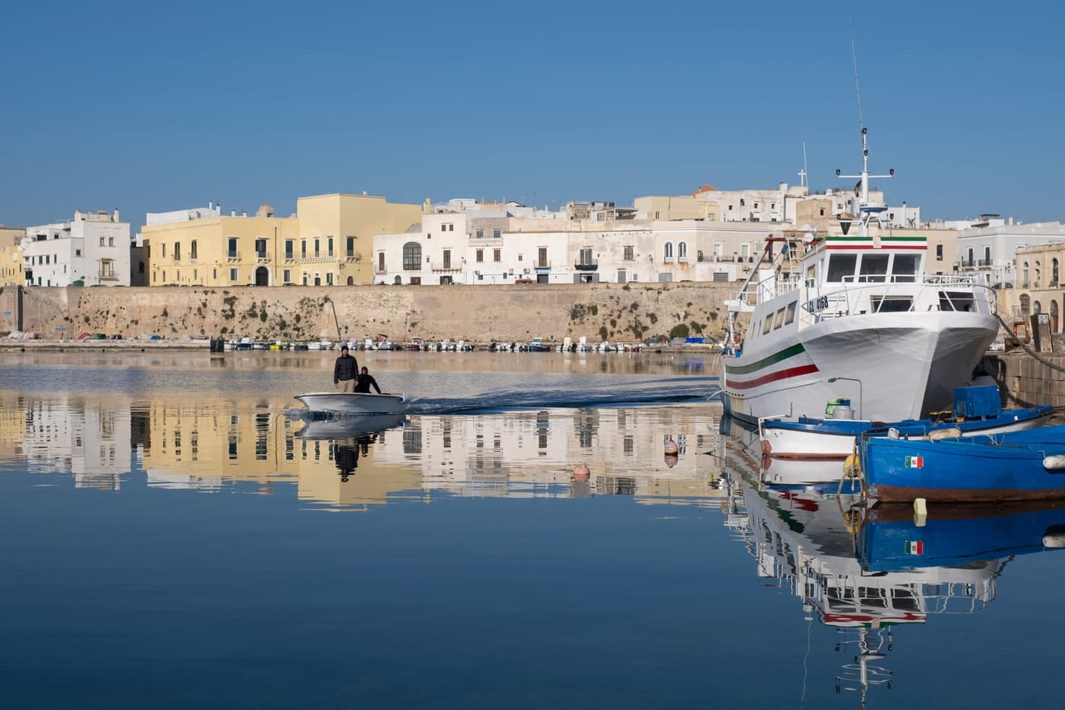 Gallipoli's old town across calm harbor water: white and pale-yellow houses on a long stone sea wall, mirrored in the water, with fishing boats moored at right