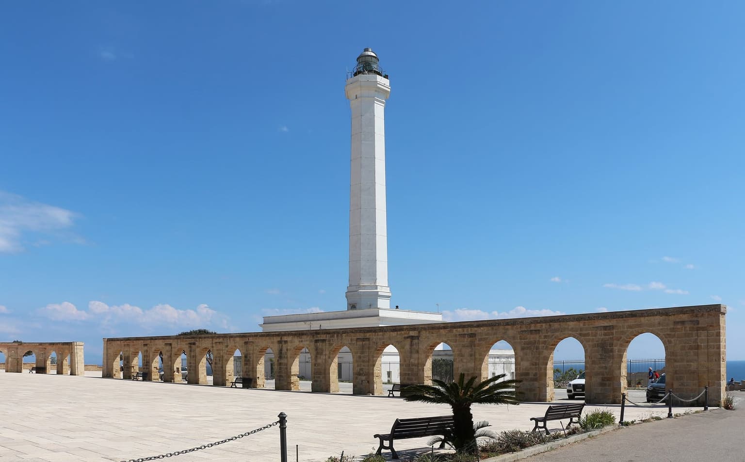 The white lighthouse at Santa Maria di Leuca behind a long arcade of pale stone arches, with a glimpse of blue sea at right