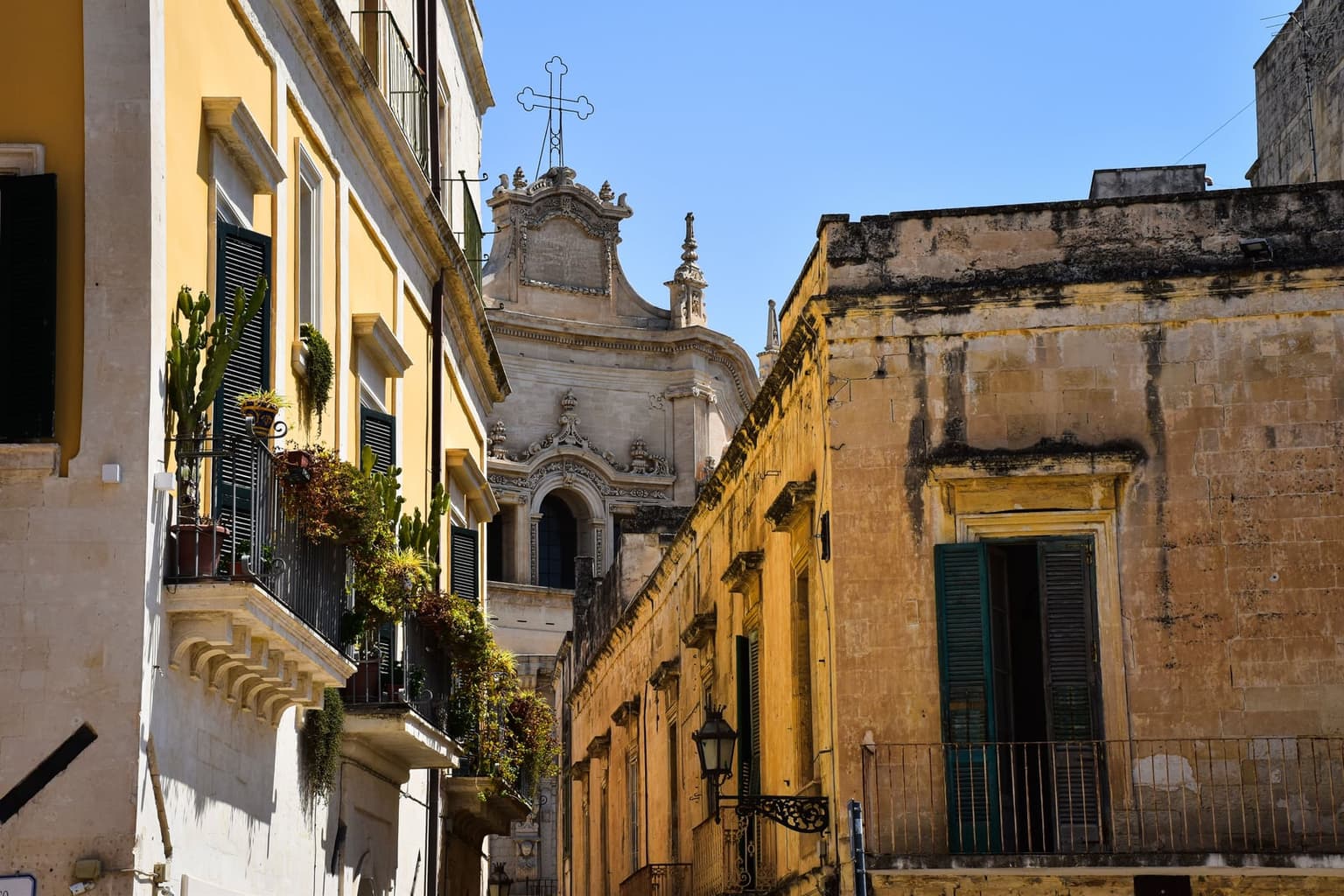 A narrow street in Lecce's old town: pale-stone and yellow houses with carved stone balconies and potted plants, green shutters, and the carved top of a baroque church at the end