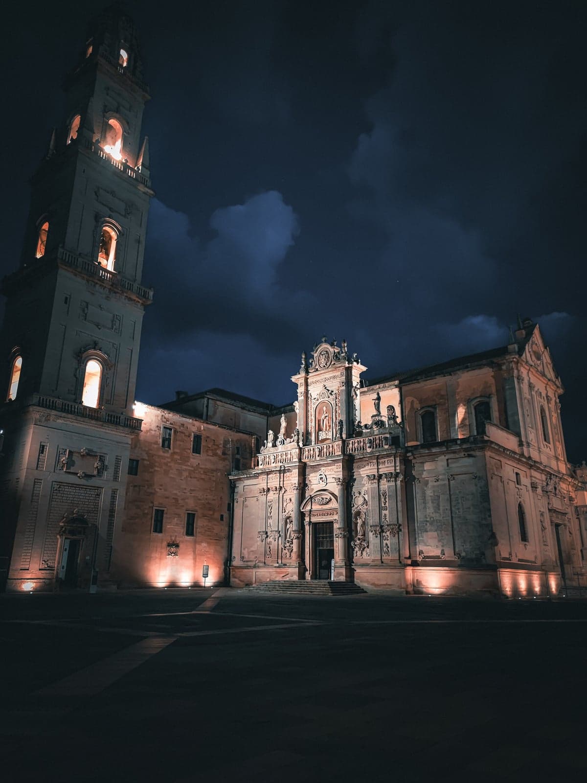 Piazza del Duomo in Lecce at night: the bell tower with its windows lit and the cathedral's carved facade in warm floodlight above the empty square