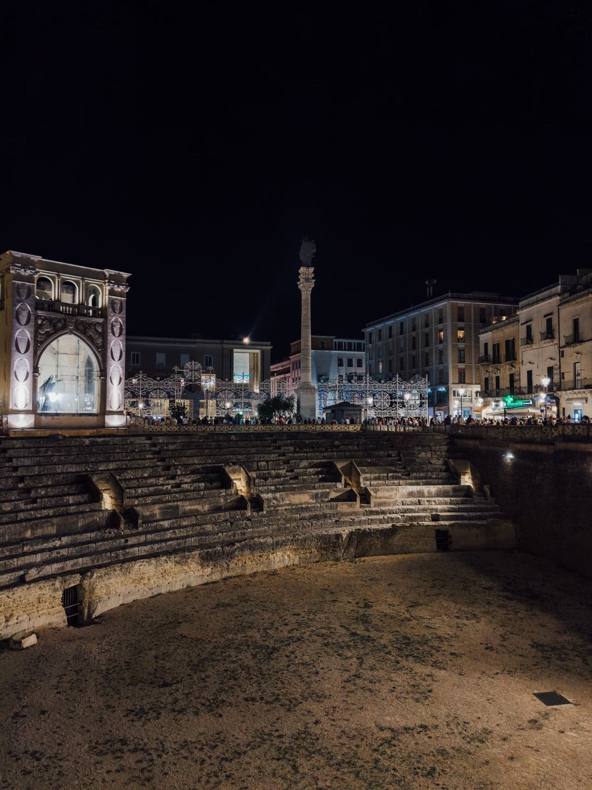 The Roman amphitheater in Piazza Sant'Oronzo at night, its stone tiers below street level, with the floodlit Sedile, the column of Sant'Oronzo, and crowds along the railing above