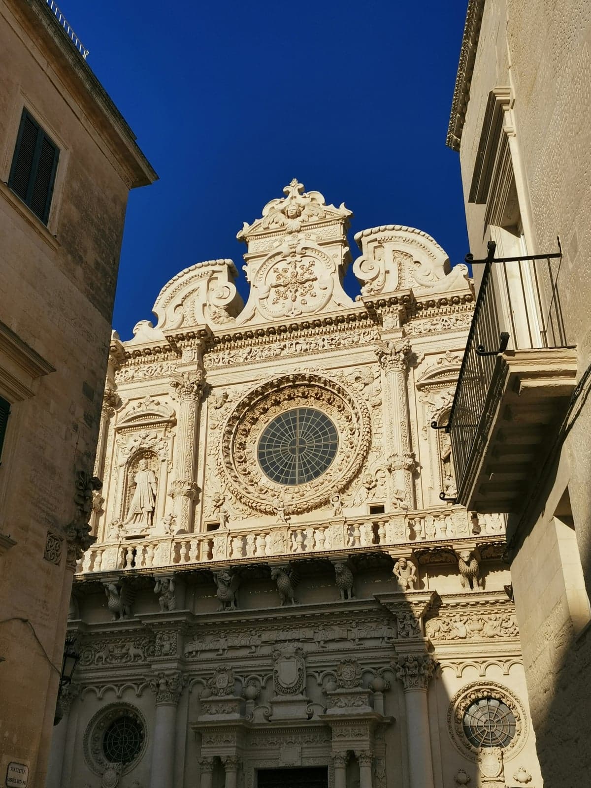 The facade of the Basilica di Santa Croce in Lecce: the round rose window ringed with carved garlands and the long balcony held up by carved figures and animals, against a blue sky