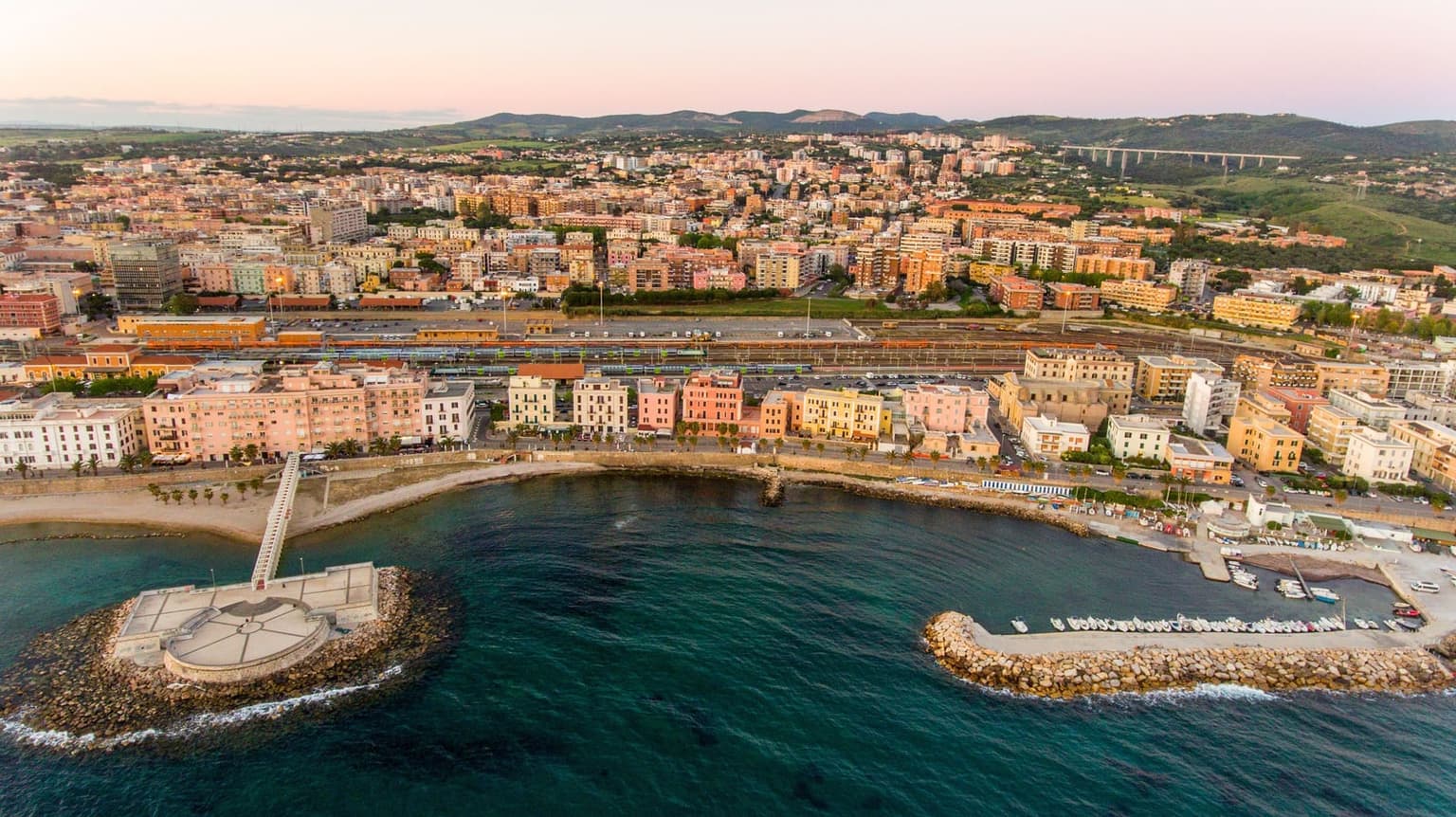 Aerial view of the Civitavecchia seafront at sunset, with a round platform on a rock pier, a curving promenade lined with pastel buildings, a small boat harbor behind a breakwater, and railway tracks just behind the waterfront