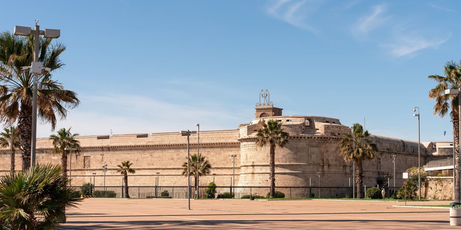 The sand-colored stone walls and round corner bastion of Forte Michelangelo in Civitavecchia, seen across a paved square lined with palm trees