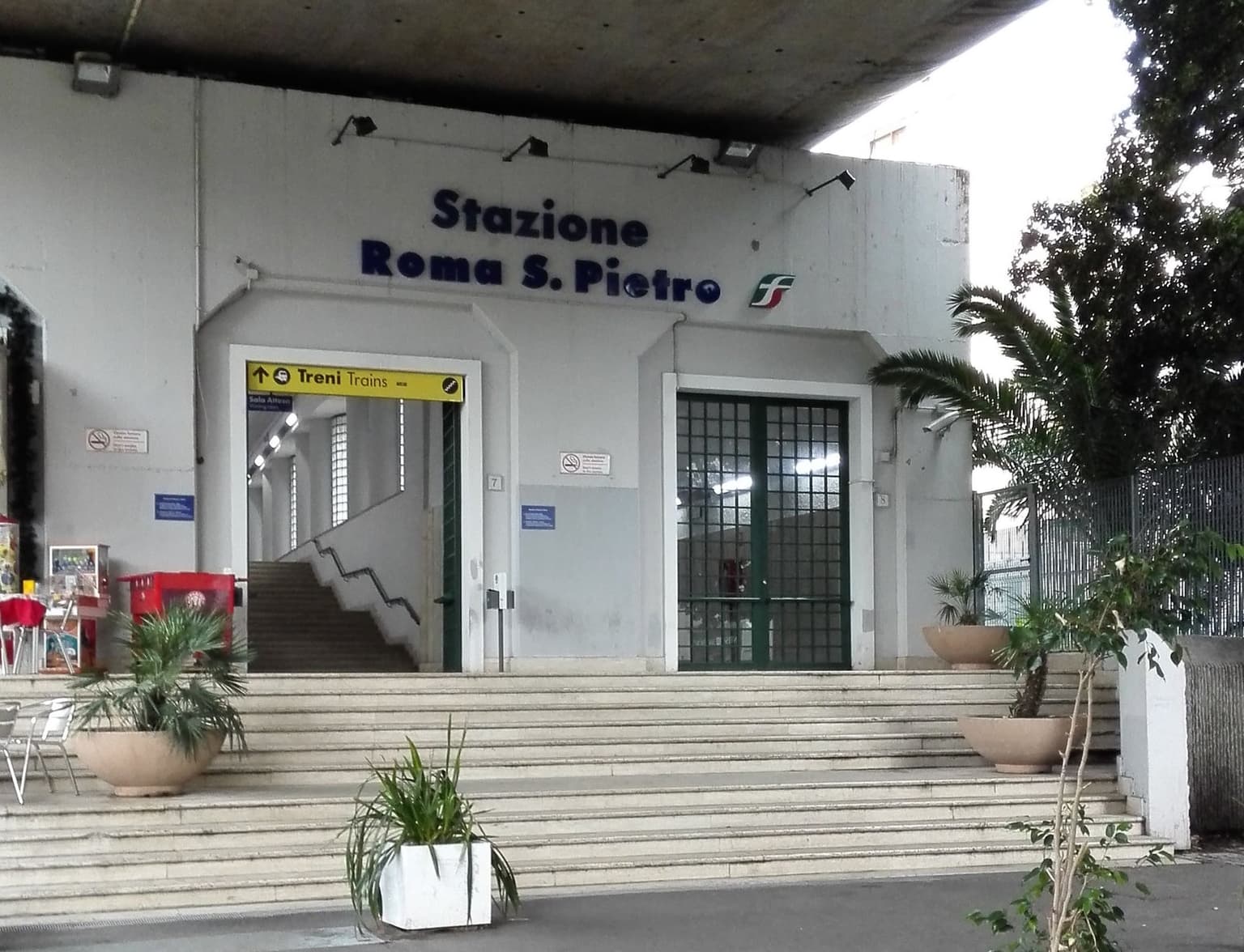Entrance of Roma San Pietro station under a concrete road overpass, with the station name above the doors and a yellow Treni Trains sign over the stairs