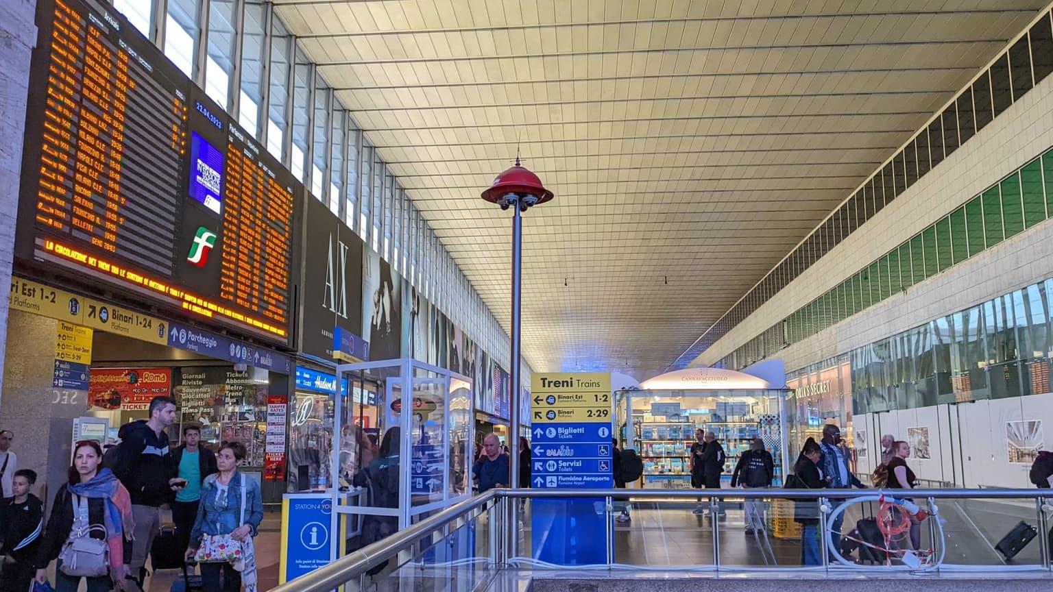 Main concourse of Roma Termini station, with a large orange departures board on the left and a blue sign pointing to the platforms and Fiumicino Airport
