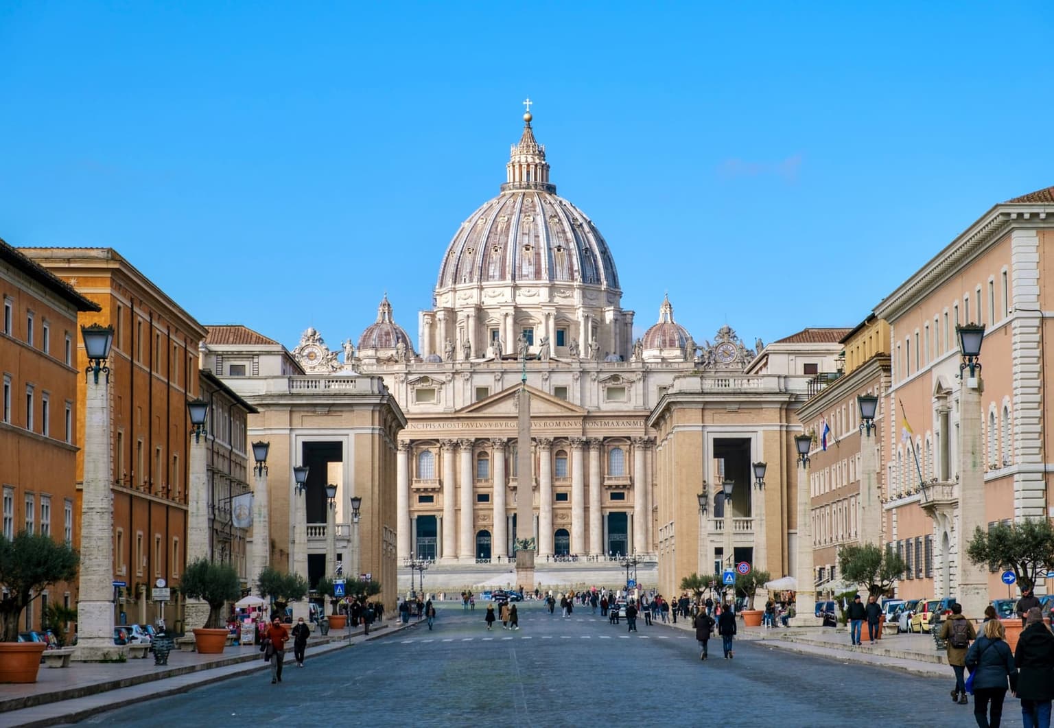 St. Peter's Basilica at the end of Via della Conciliazione in Rome, its dome and facade framed by the avenue's stone lamp columns, with the obelisk of St. Peter's Square in front