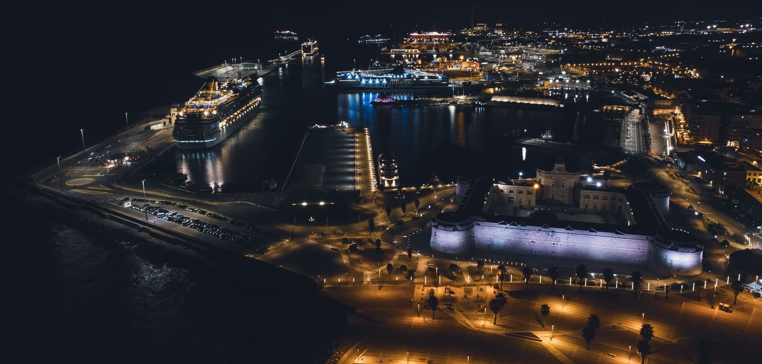 Night aerial view of Civitavecchia port, with a large cruise ship lit up at a quay, ferries and more ships across the basin, and the floodlit walls of Forte Michelangelo at the lower right