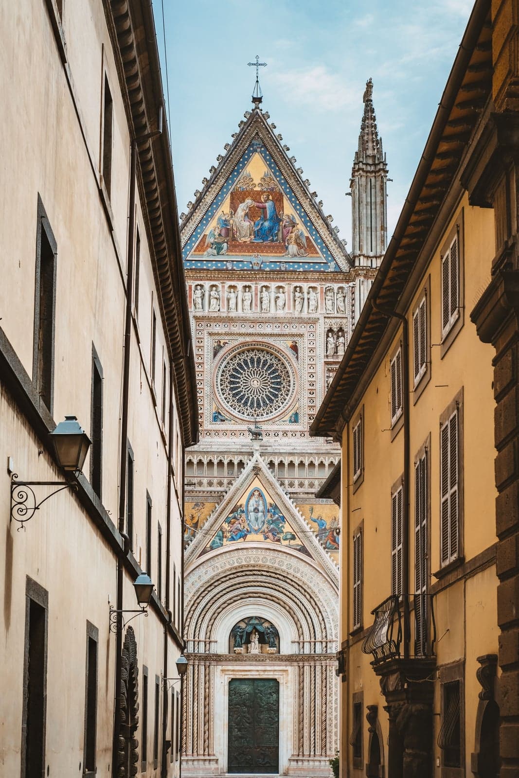 The front of Orvieto Cathedral seen down a narrow street between pale buildings, its gold mosaic gables, round rose window, and bronze central door filling the gap