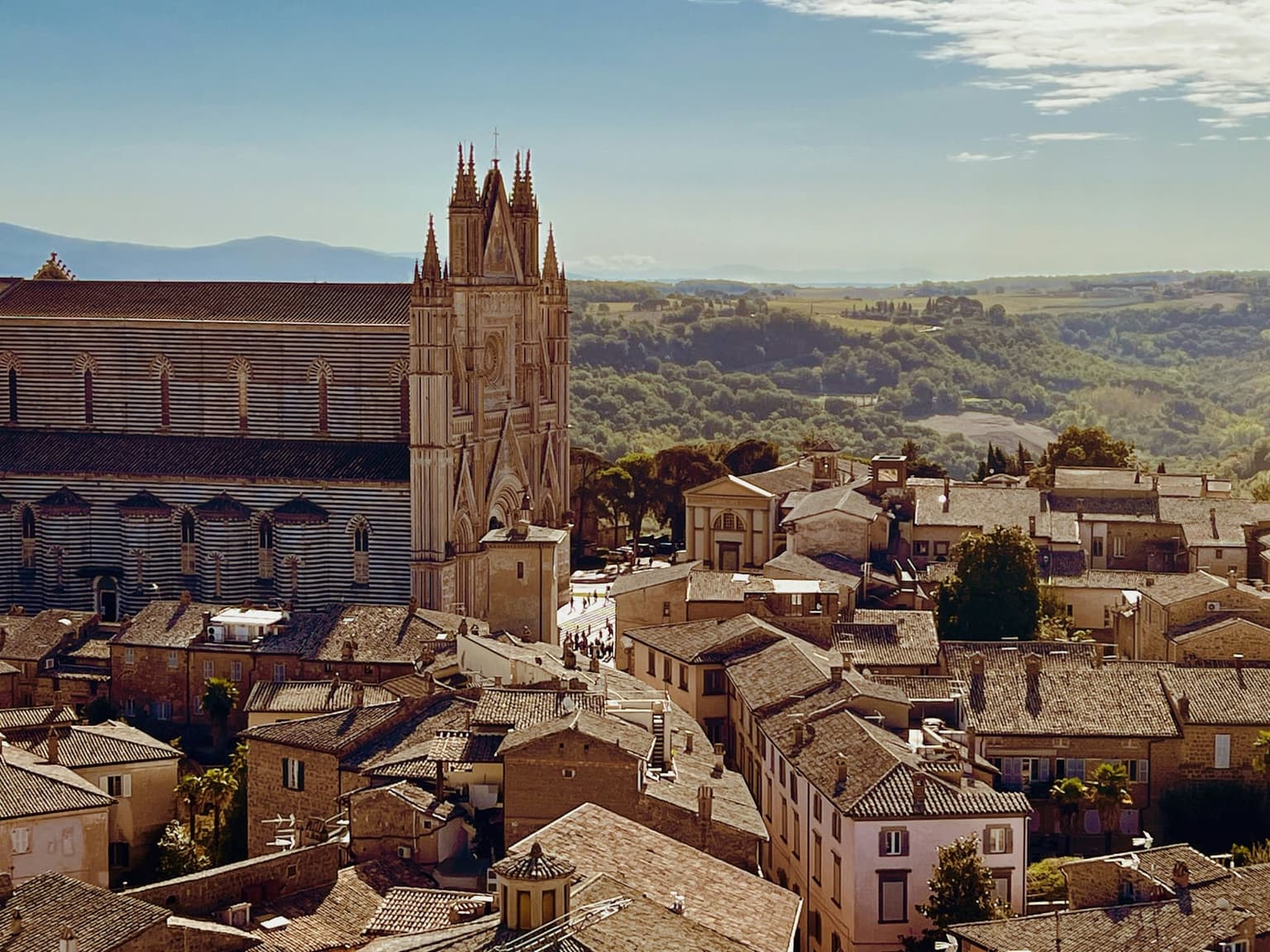 Orvieto Cathedral's striped stone flank and pointed Gothic front rising above the tiled rooftops of Orvieto's old town, with green hills and farmland stretching behind
