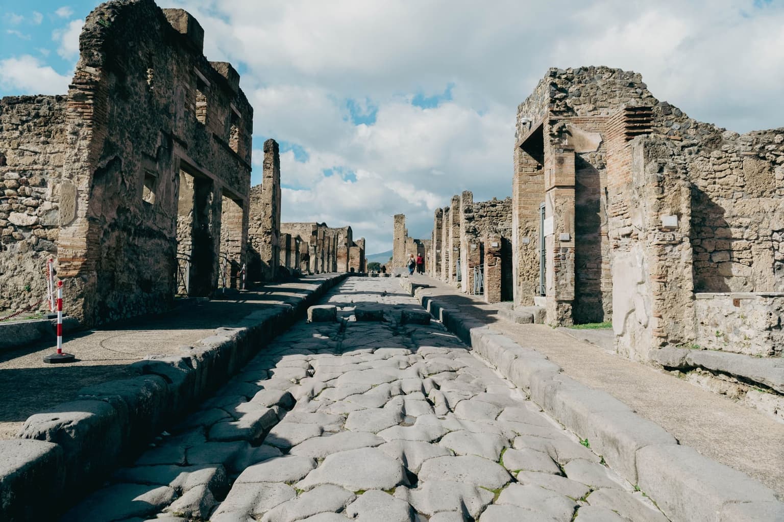 A long paved street inside ancient Pompeii, its large worn lava stones running between raised sidewalks and a row of stepping stones, with ruined stone and brick houses on both sides and Vesuvius faint at the far end