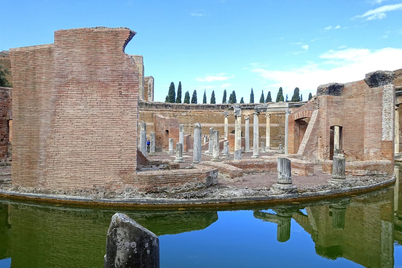 The round Maritime Theater at Hadrian's Villa near Tivoli, brick walls and fluted columns on a small island ringed by a still moat that mirrors the ruins, with cypresses along the skyline
