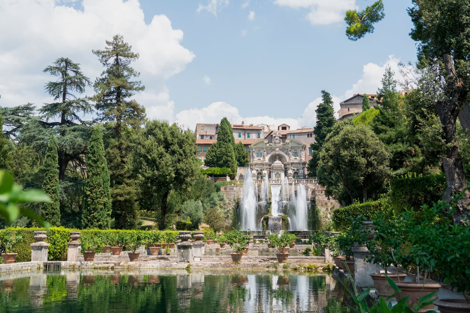 The fishponds of Villa d'Este in Tivoli, with tall fountain jets rising behind them below the ornate Organ Fountain, the town's houses on the hill above, and cypresses on both sides
