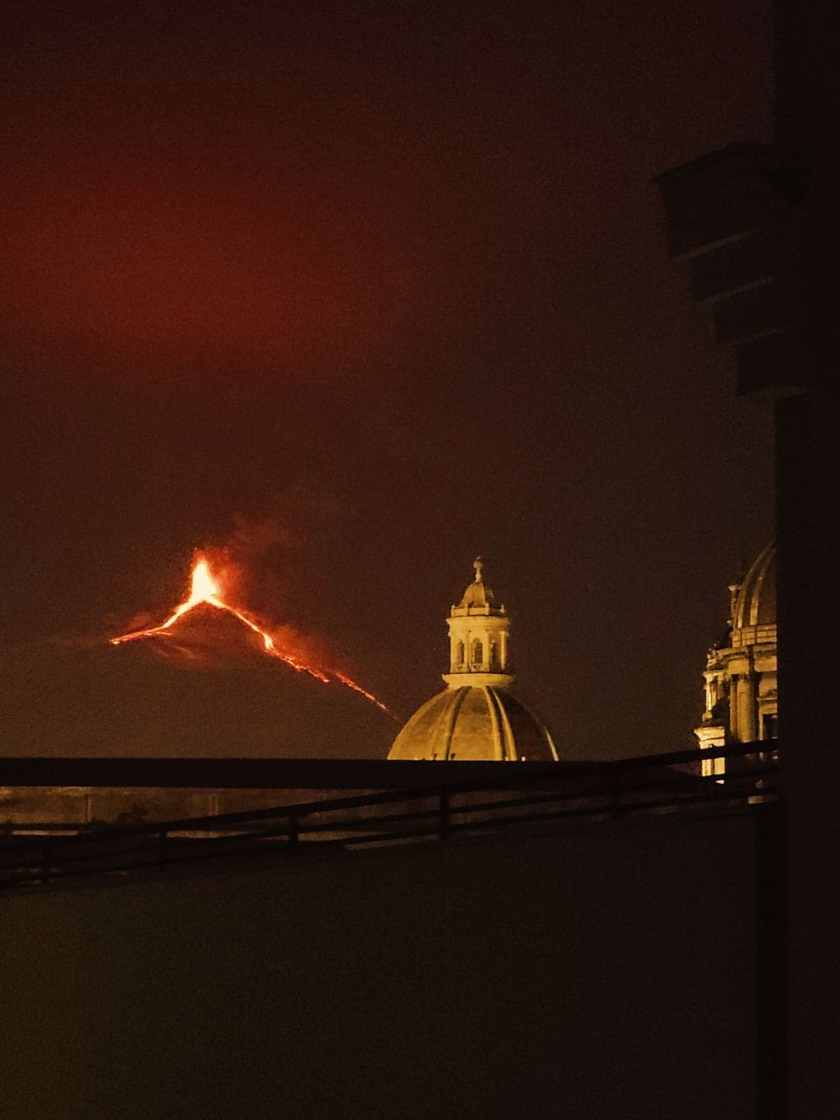 Mount Etna erupting at night, a bright orange lava fountain and glowing lava flow visible on the distant slope against a dark red sky, seen past the floodlit dome and tower of a church in the foreground, with a rooftop railing at the bottom of the frame.