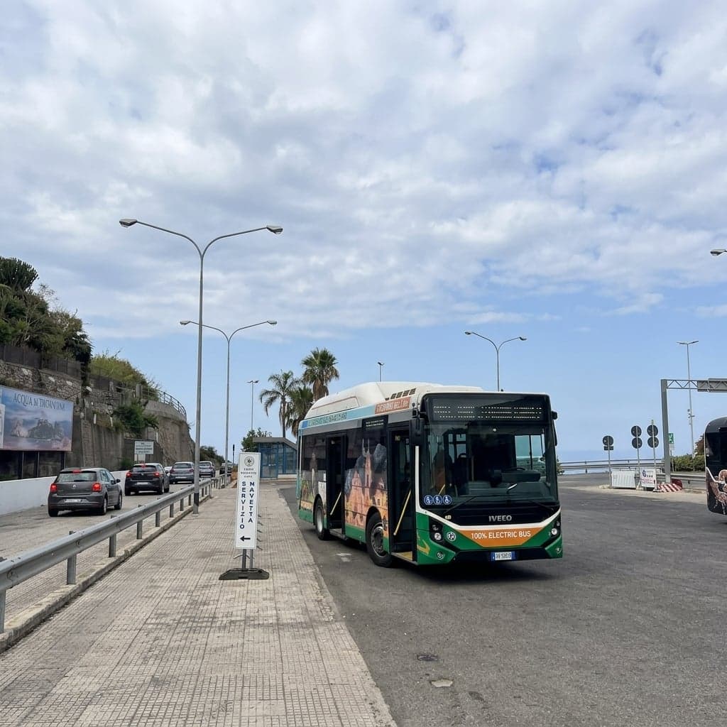 A green-and-white ASM Taormina electric shuttle bus, its side painted with a mural of Taormina's skyline landmarks, parked at the Lumbi shuttle stop beside a small blue Servizio Navetta sign, with palm trees, parked cars along a guardrail, and another coach at the edge of the frame.