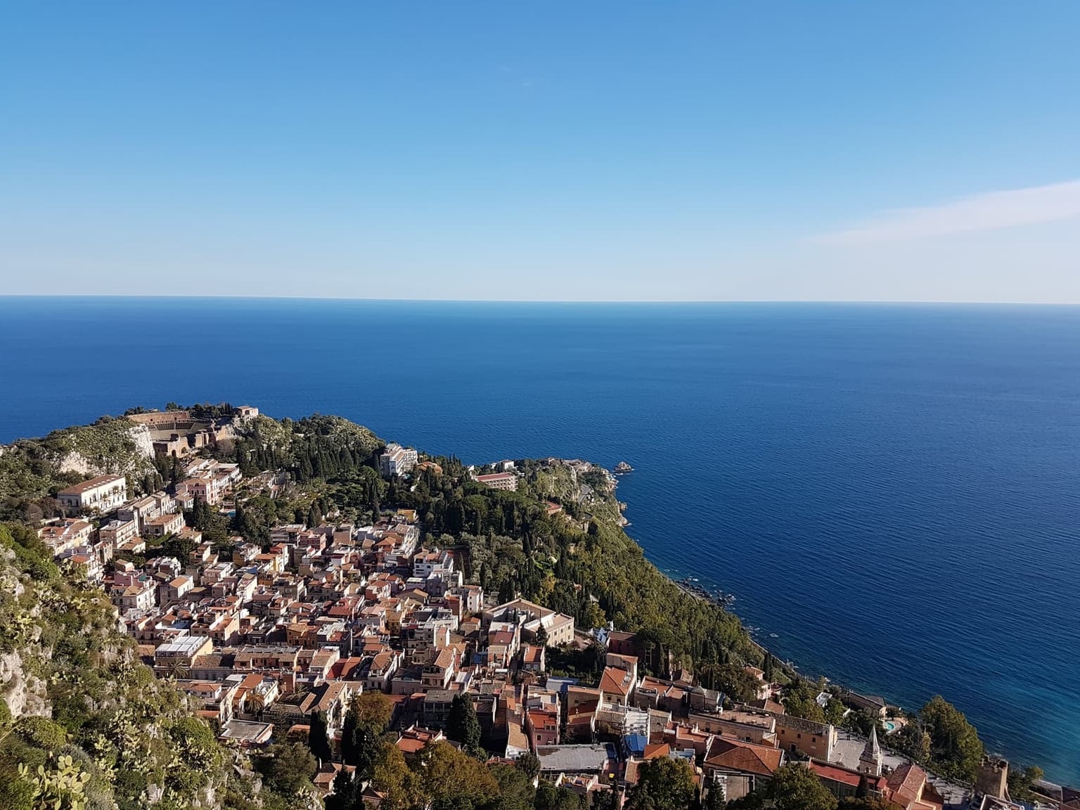 Aerial view of Taormina's old town clustered along a green ridge above the Ionian Sea, the ruins of the ancient Greek theater visible on the headland at upper left, terracotta rooftops descending the hillside toward the cypress-lined coast, open blue sea filling the horizon under a clear sky.