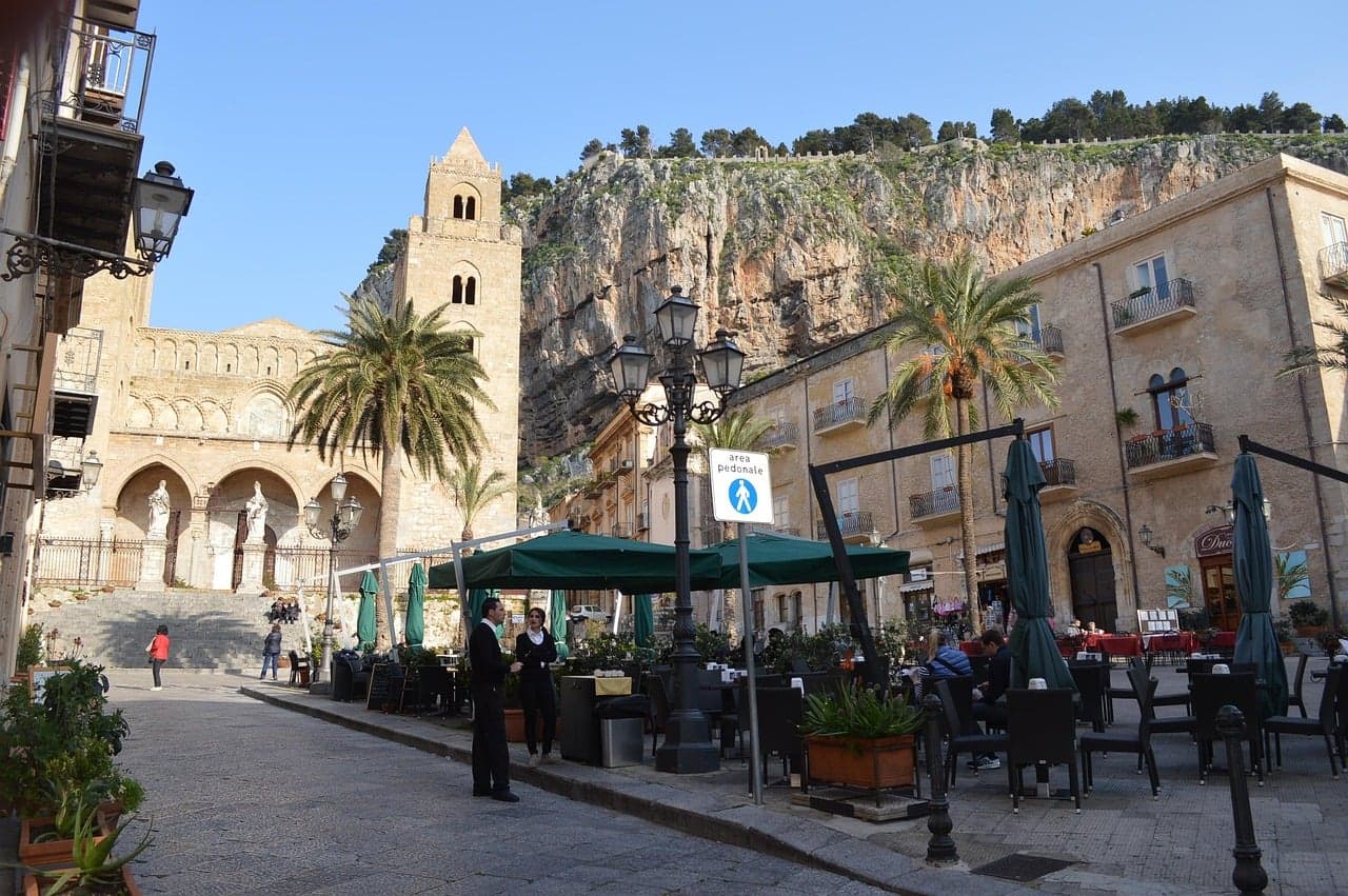 Piazza Duomo in Cefalù, the cathedral's arched porch and bell tower beside palms and café umbrellas, with the limestone cliff of La Rocca rising behind