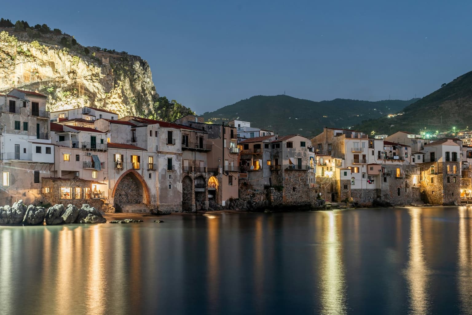 Cefalù's old waterfront at dusk, lit houses and a pointed arch reflected in calm water under the floodlit cliff of La Rocca
