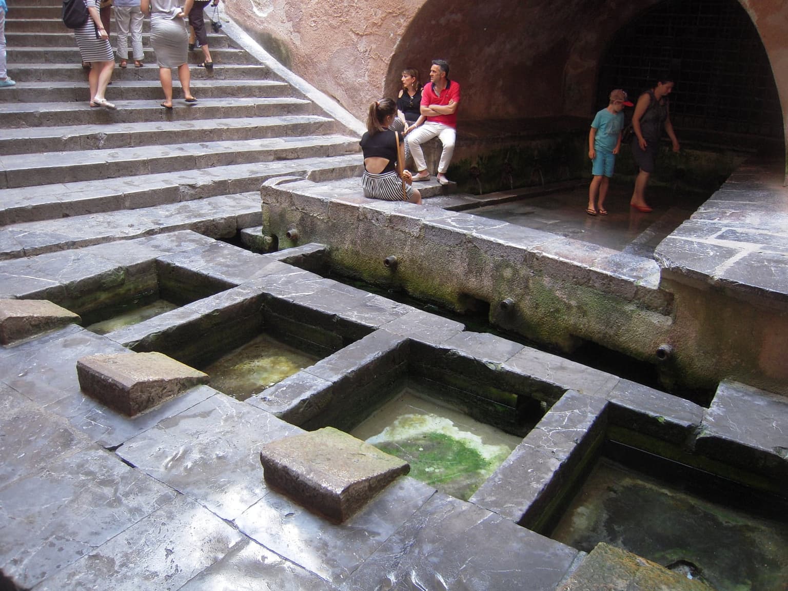 The medieval Lavatoio in Cefalù, stone basins of shallow water below a wide staircase and a vaulted arch, with visitors on the steps