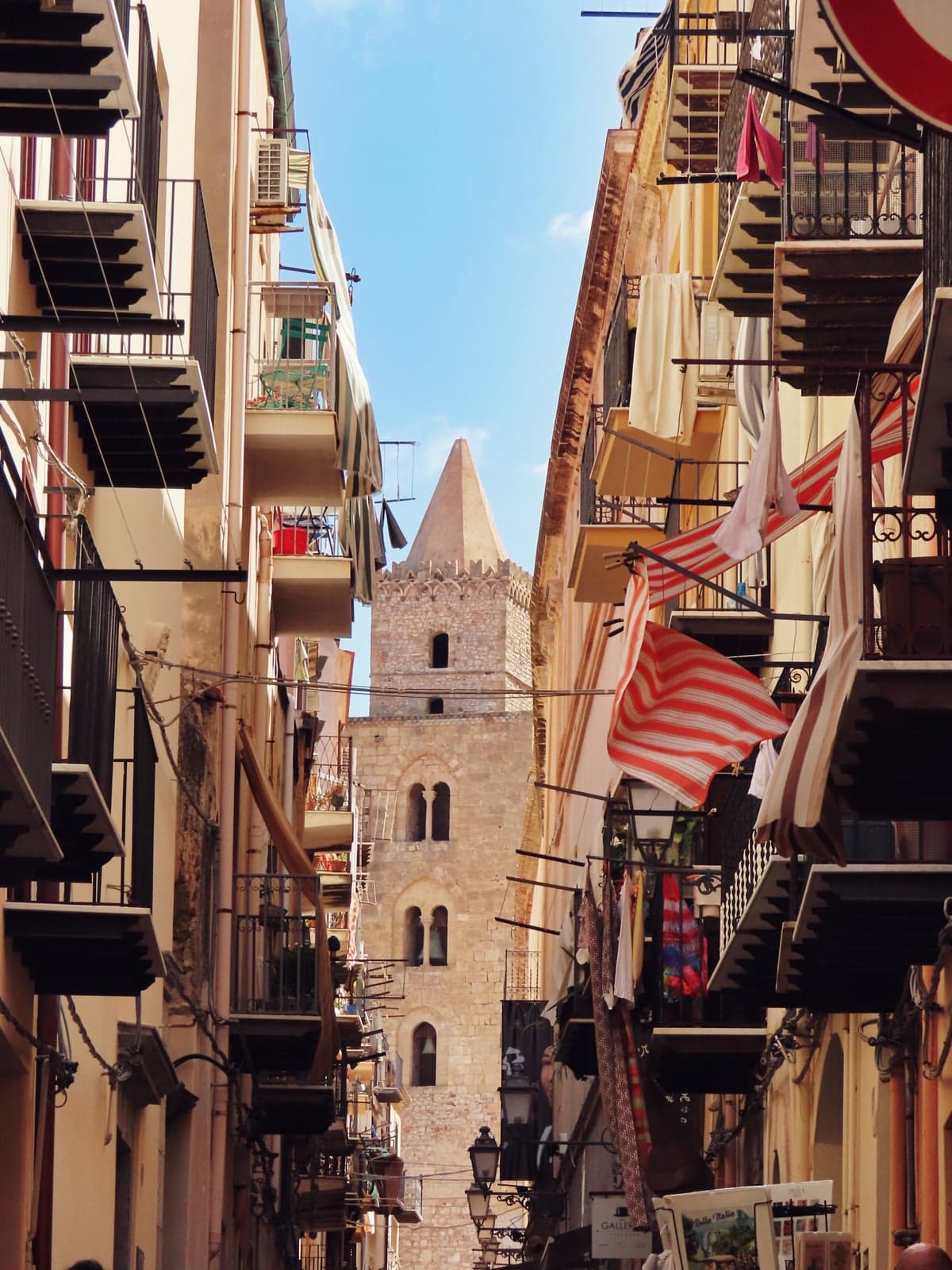 A narrow old-town lane in Cefalù with iron balconies, laundry, and striped awnings, the cathedral's square tower at its far end