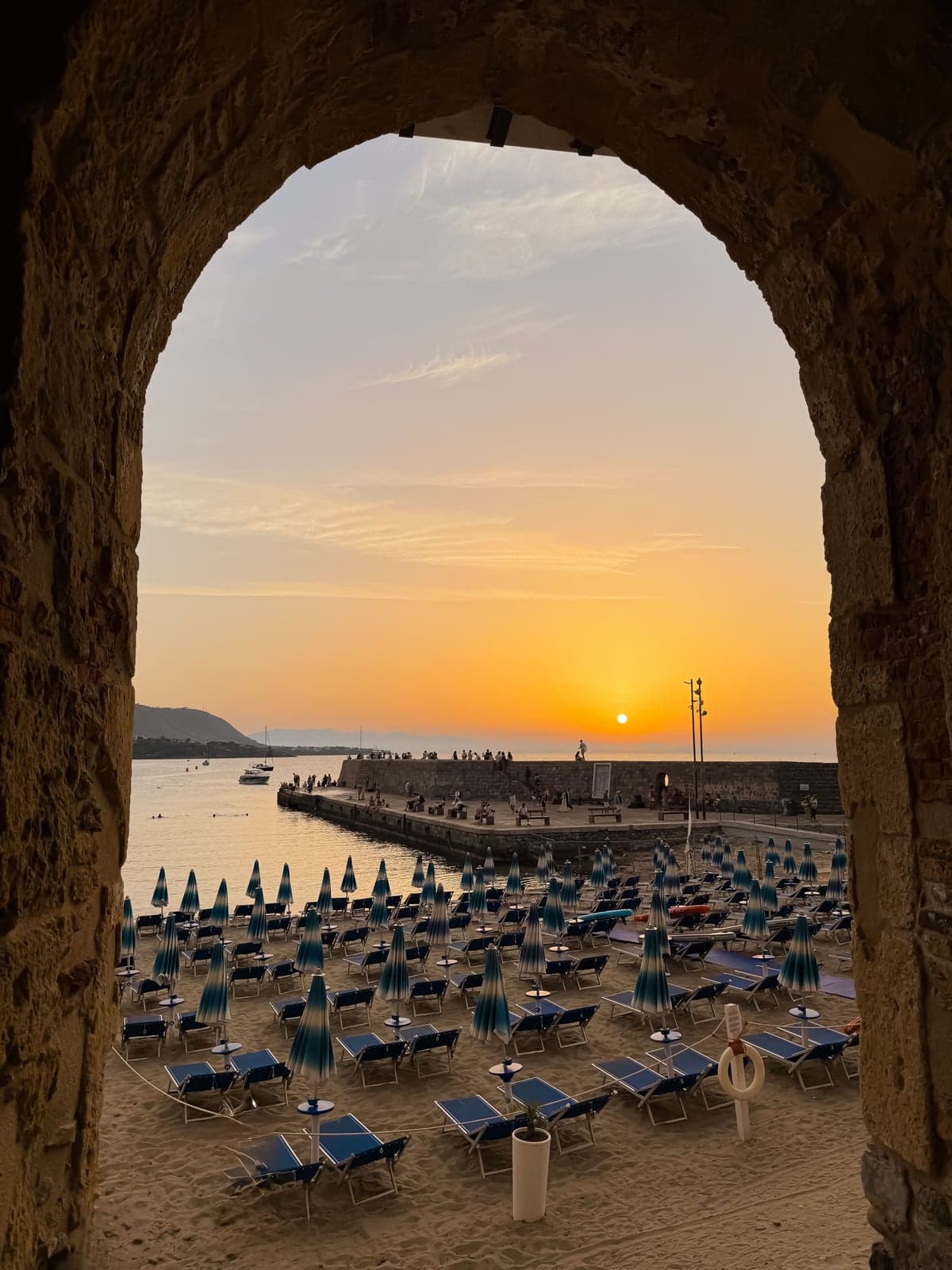 Sunset through the arch of Porta Pescara in Cefalù, looking out at the old pier and rows of closed beach umbrellas
