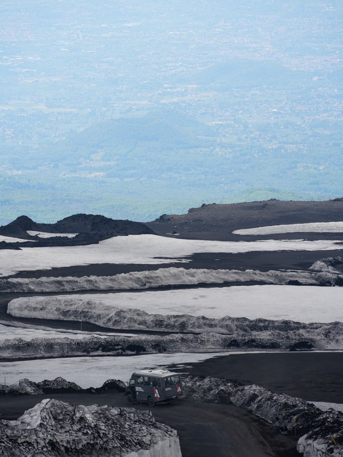 A gray 4x4 bus marked Funivia dell'Etna crosses a black-and-white ash-and-snow slope high on the volcano, with the hazy coastal plain visible far below.