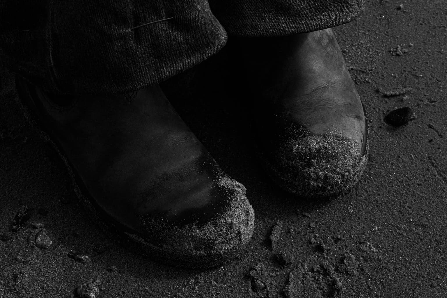 Close-up black-and-white shot of scuffed boots dusted in fine black grit, standing on dark volcanic-looking ground.